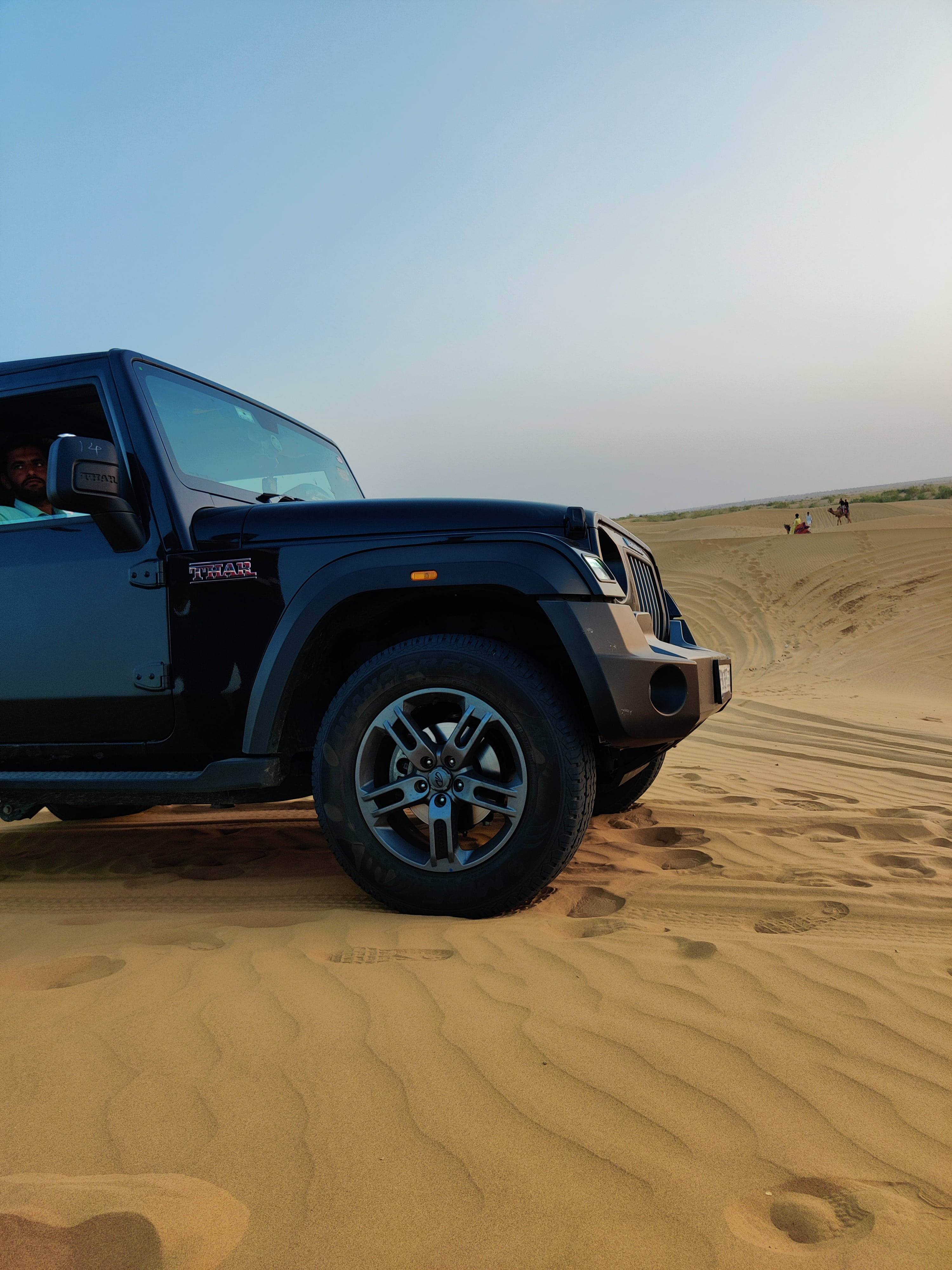 手機 桌布 壁紙 — 一輛黑色Jeep Wrangler在晴朗藍天下駛過金色沙漠沙丘，遠處可見人影。