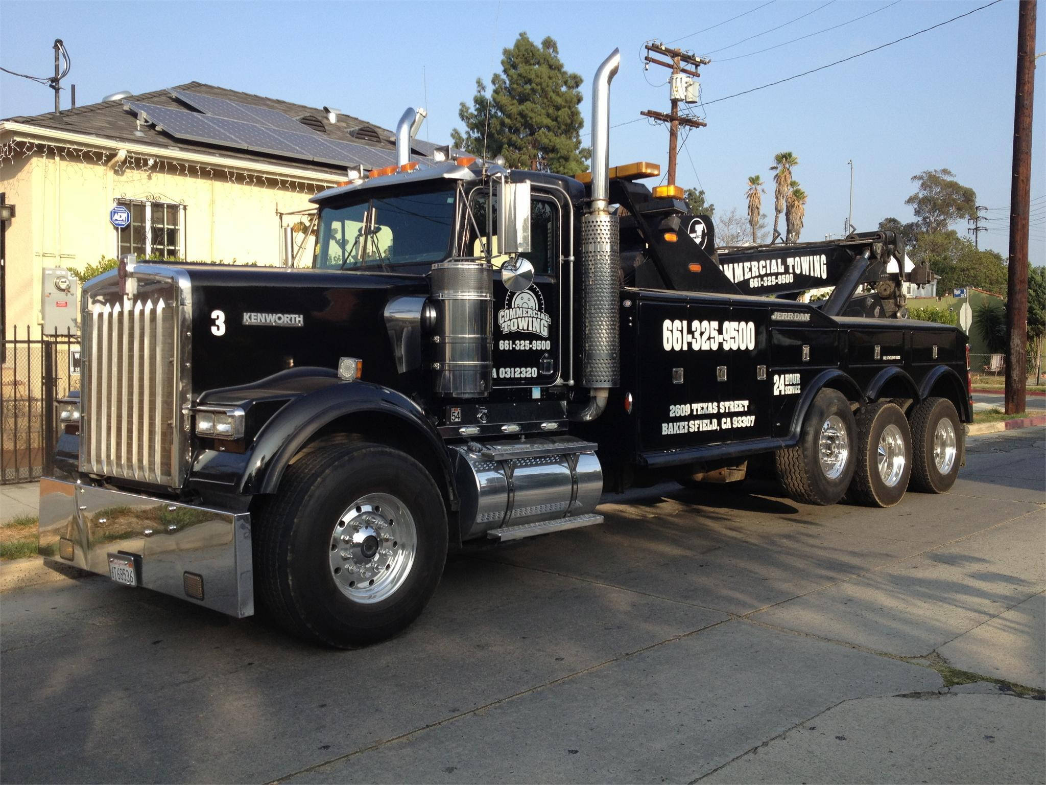 Kenworth W900 桌布 壁紙 — 一輛黑色Kenworth拖車，帶有鍍鉻裝飾和可見的拖車設備，停放在陽光明媚的街道上，旁邊是一棟裝有太陽能電池板的房子。