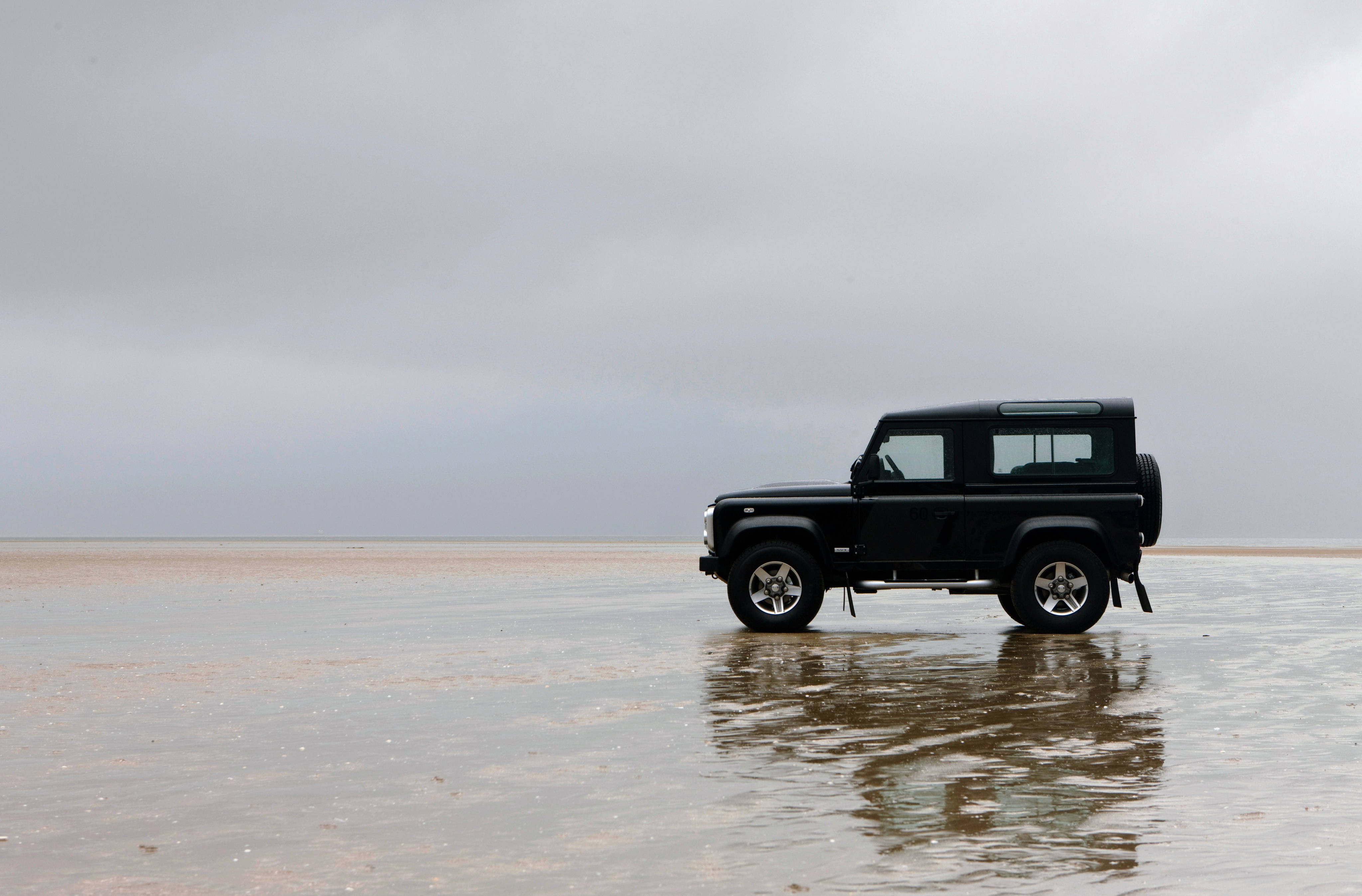 land rover iPhone 桌布 壁紙 — 一輛黑色Land Rover Defender獨自停在一片廣闊的濕沙灘上，天空多雲，倒影在淺水中閃爍。