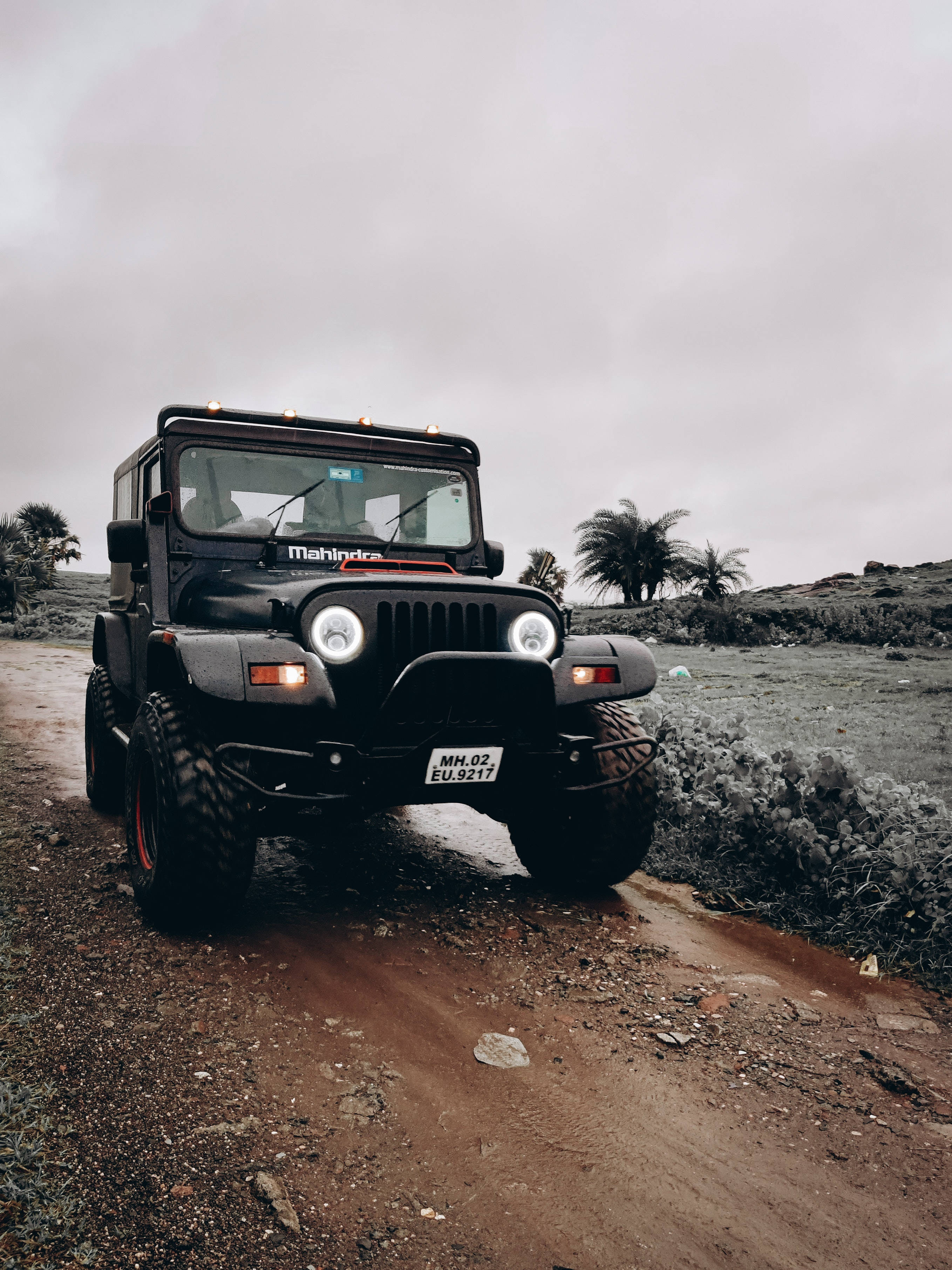 人像動態 桌布 壁紙 — 一輛黑色Mahindra Thar SUV亮著車頭燈，在泥濘的越野小徑上行駛，背景是陰天和棕櫚樹。
