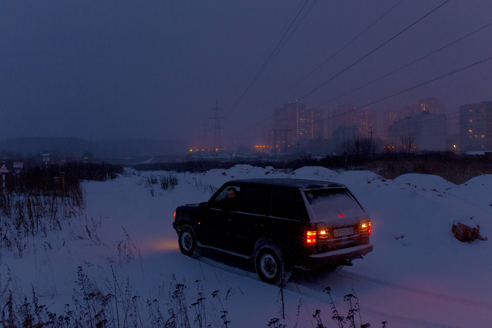 夜空 桌布 壁紙 — 一輛黑色SUV在黃昏時分駛過雪地，遠處的城市燈光在黑暗的天空下閃爍。