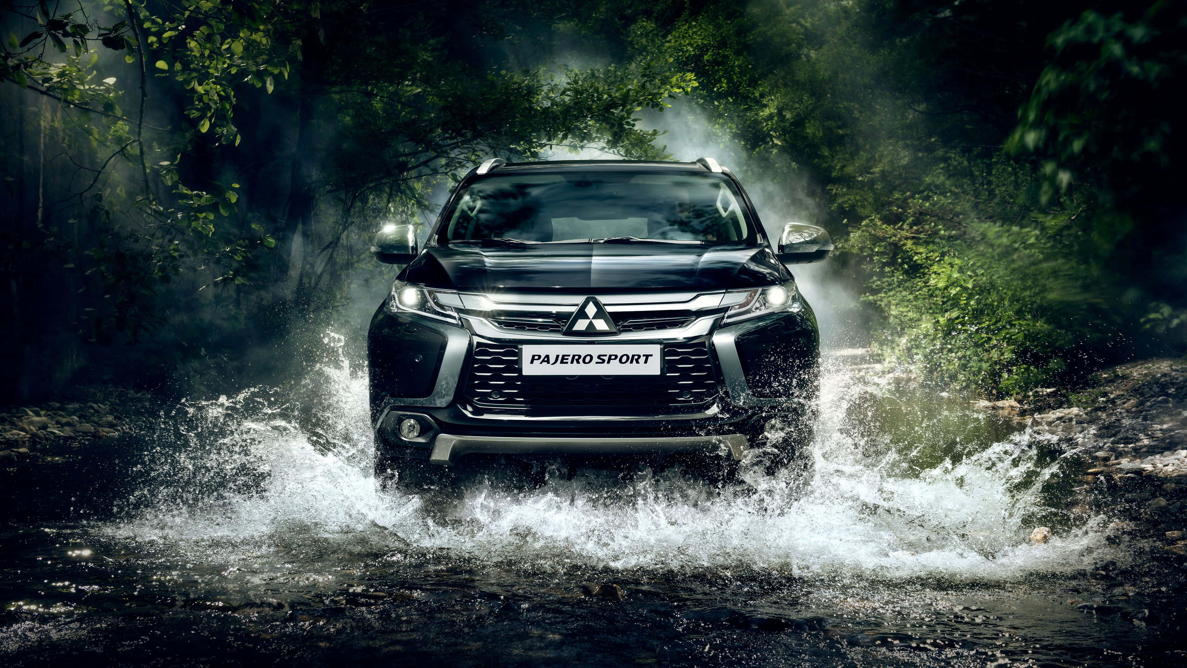 Mitsubishi Pajero 桌布 壁紙 — 一輛黑色三菱帕杰羅運動版SUV在茂密的綠樹下，強勁地駛過泥濘的森林溪流，戲劇性地濺起水花。