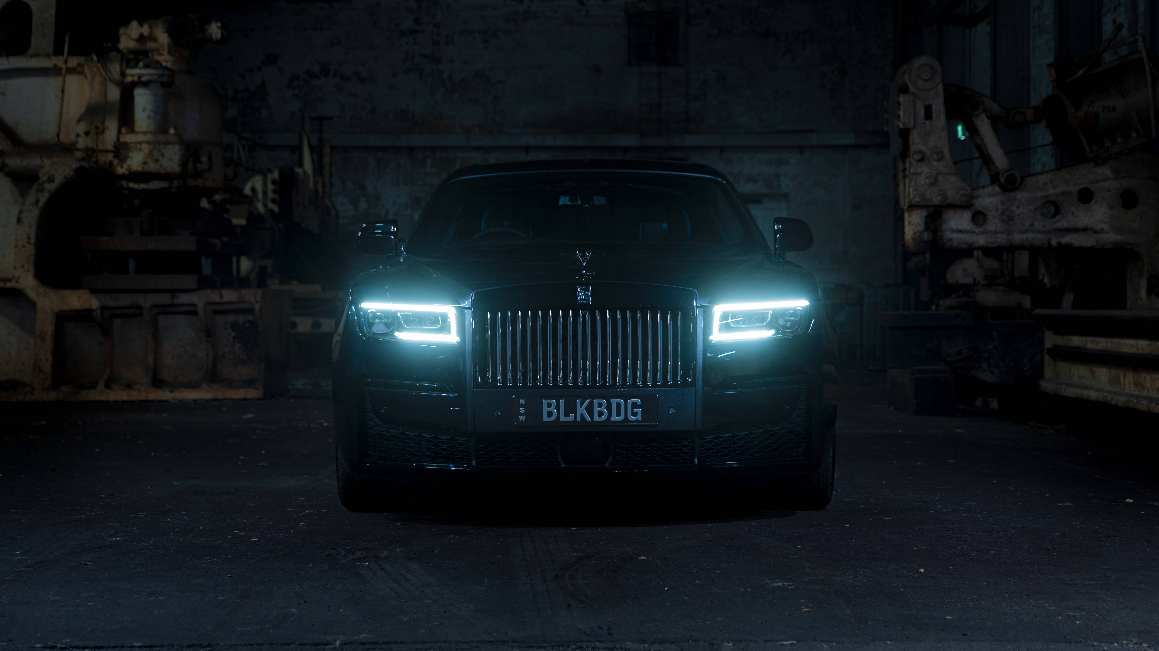 Royce 桌布 壁紙 — 一輛黑色勞斯萊斯 Ghost 在昏暗的工業倉庫中閃爍著銳利的車頭燈，將強光投射在生鏽的機器上。