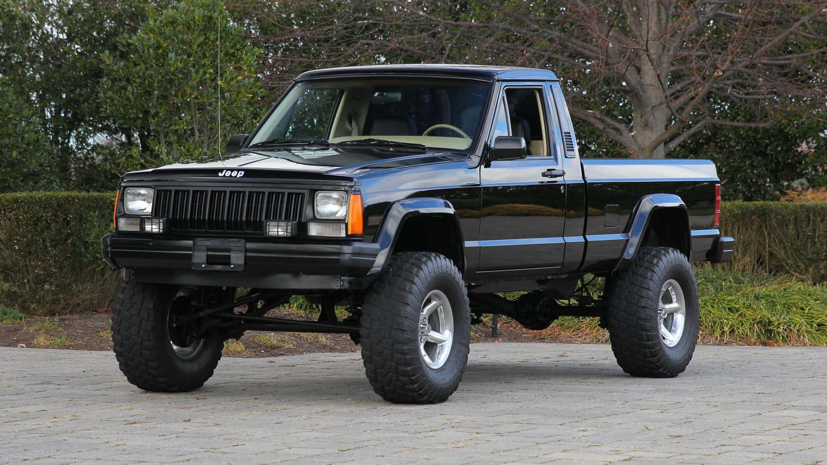 Jeep Comanche 桌布 壁紙 — 一輛黑色升高底盤的Jeep Cherokee皮卡車，配備大型越野輪胎，停在鋪砌的車道上，周圍綠樹成蔭。