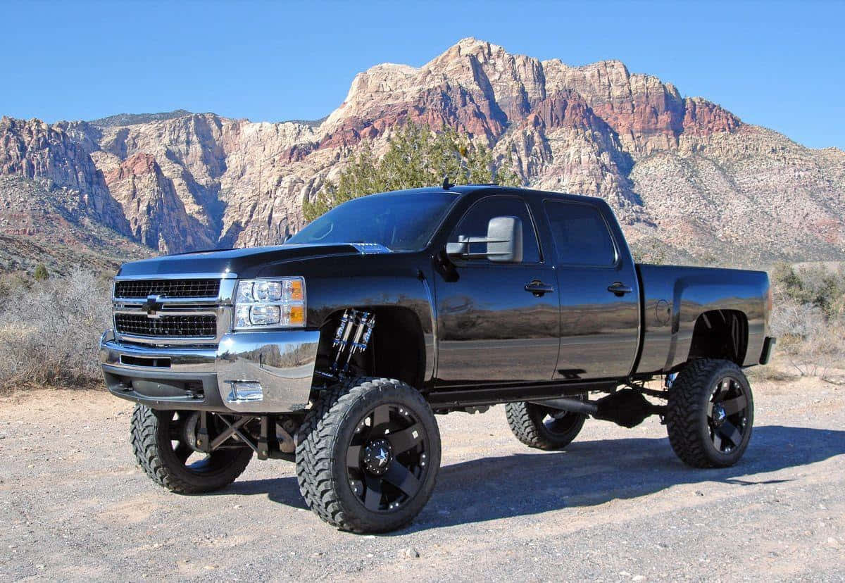 Jacked Up Trucks 桌布 壁紙 — 一輛黑色升高底盤的雪佛蘭 Silverado 皮卡車，配備大型輪胎，停泊在泥路上，前方是崎嶇的紅色岩石峽谷，天空清澈湛藍。