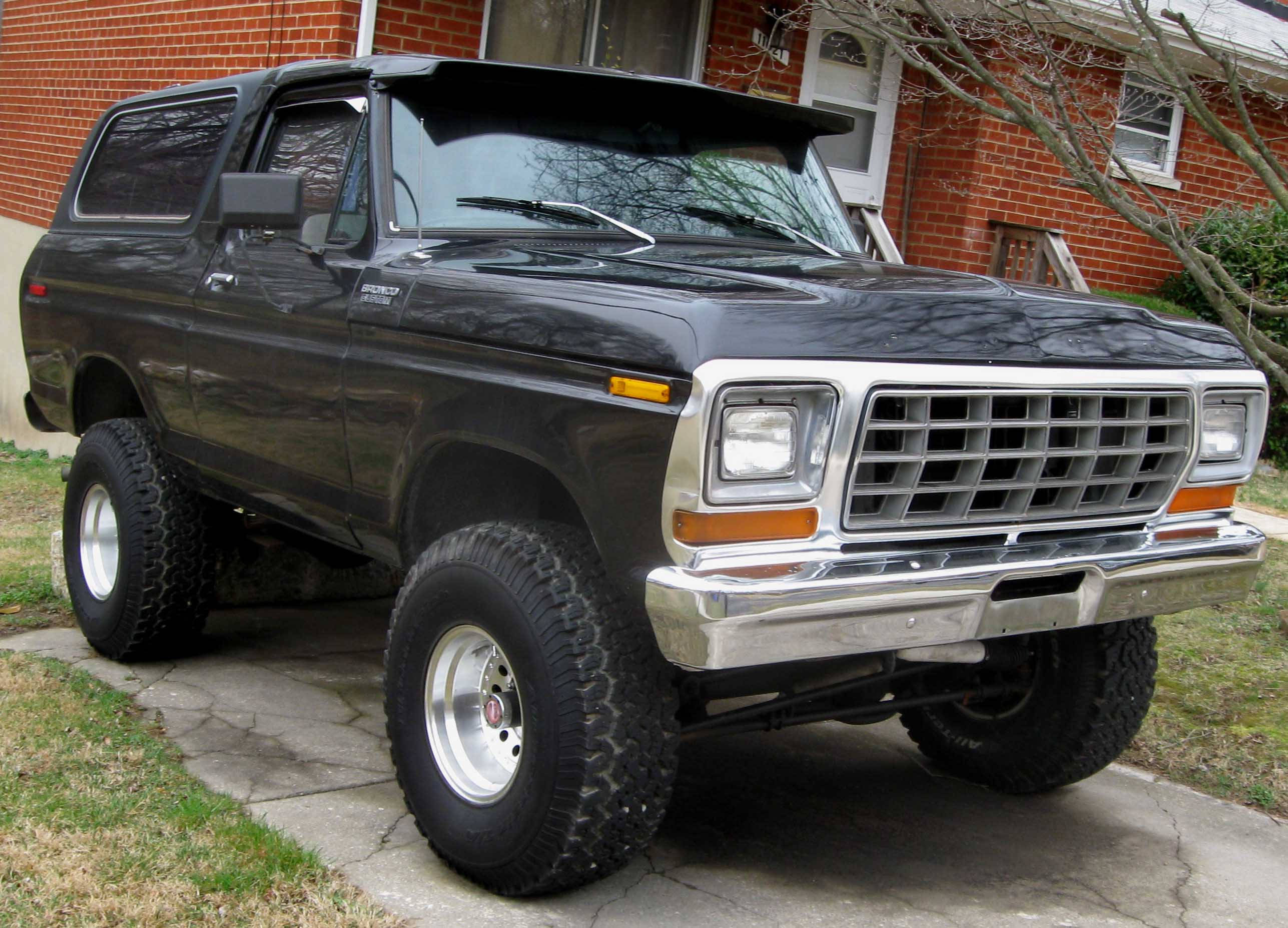 Ford Bronco 桌布 壁紙 — 一輛黑色升高版福特Bronco SUV，配備鍍鉻格柵和大輪胎，停在紅磚屋前裂開的混凝土車道上。