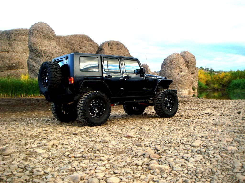 Black Jeep 桌布 壁紙 — 一輛黑色升高的Jeep Wrangler配備超大輪胎，停在岩石河岸旁，旁邊有高聳的砂岩地貌和秋葉。