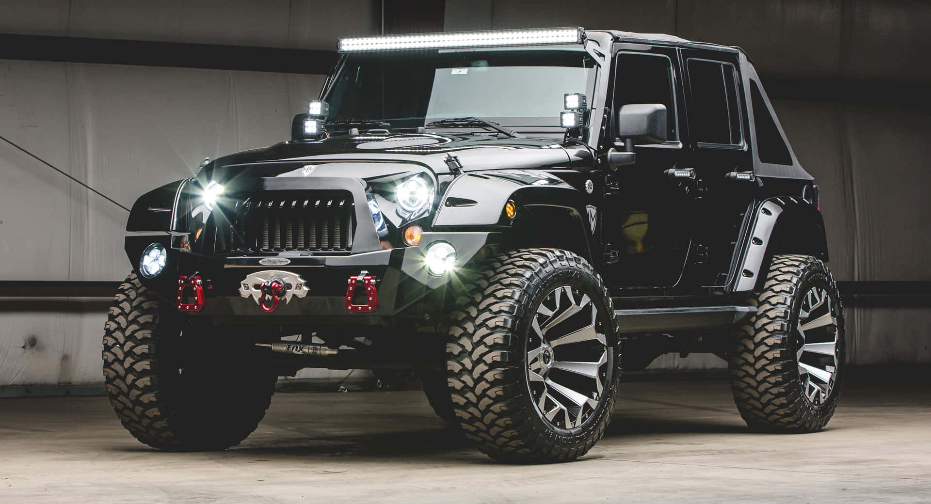 Jeep Wrangler 桌布 壁紙 — 一輛黑色升高的Jeep Wrangler，配備超大輪胎和明亮LED燈，在昏暗的車庫中顯眼地停放。