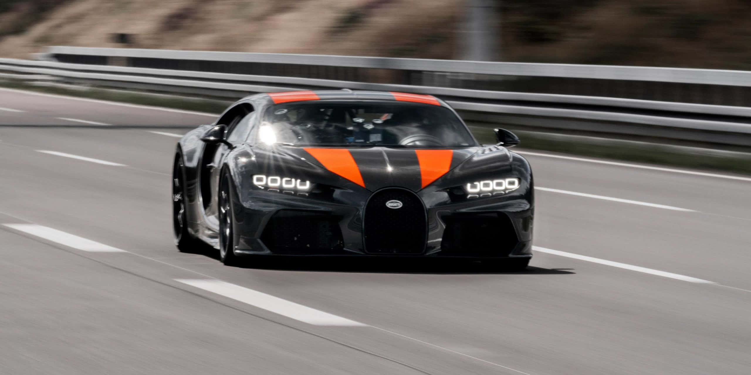 布加迪 桌布 壁紙 — 一輛黑色和橙色的Bugatti Chiron Super Sport 300+在陽光明媚的賽道上奔馳，護欄和瀝青路面呈現動態模糊效果。
