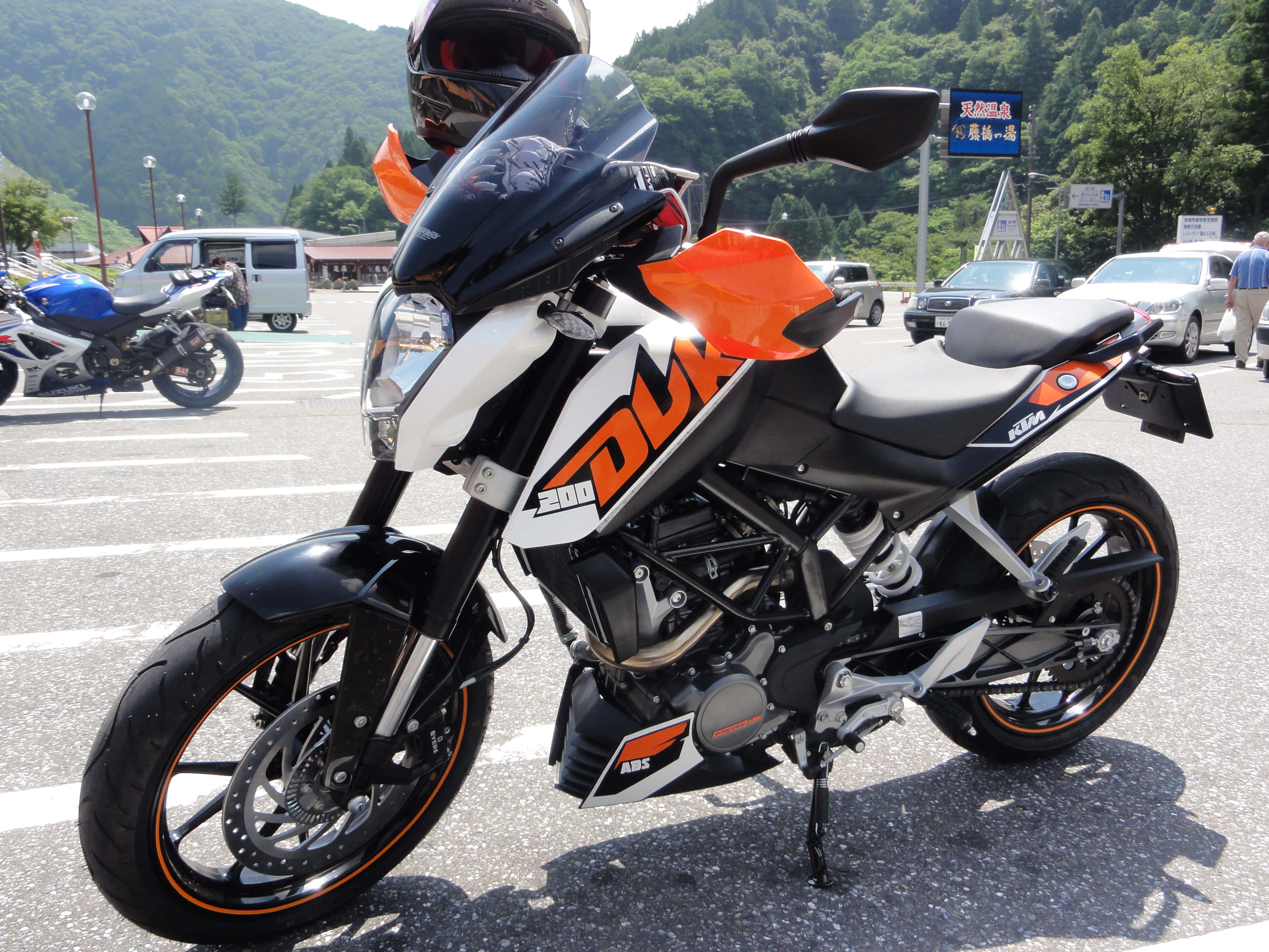 Ktm Duke 桌布 壁紙 — 一輛黑色和橙色的KTM Duke 390機車停放在陽光明媚的停車場，背景是綠色的山丘。
