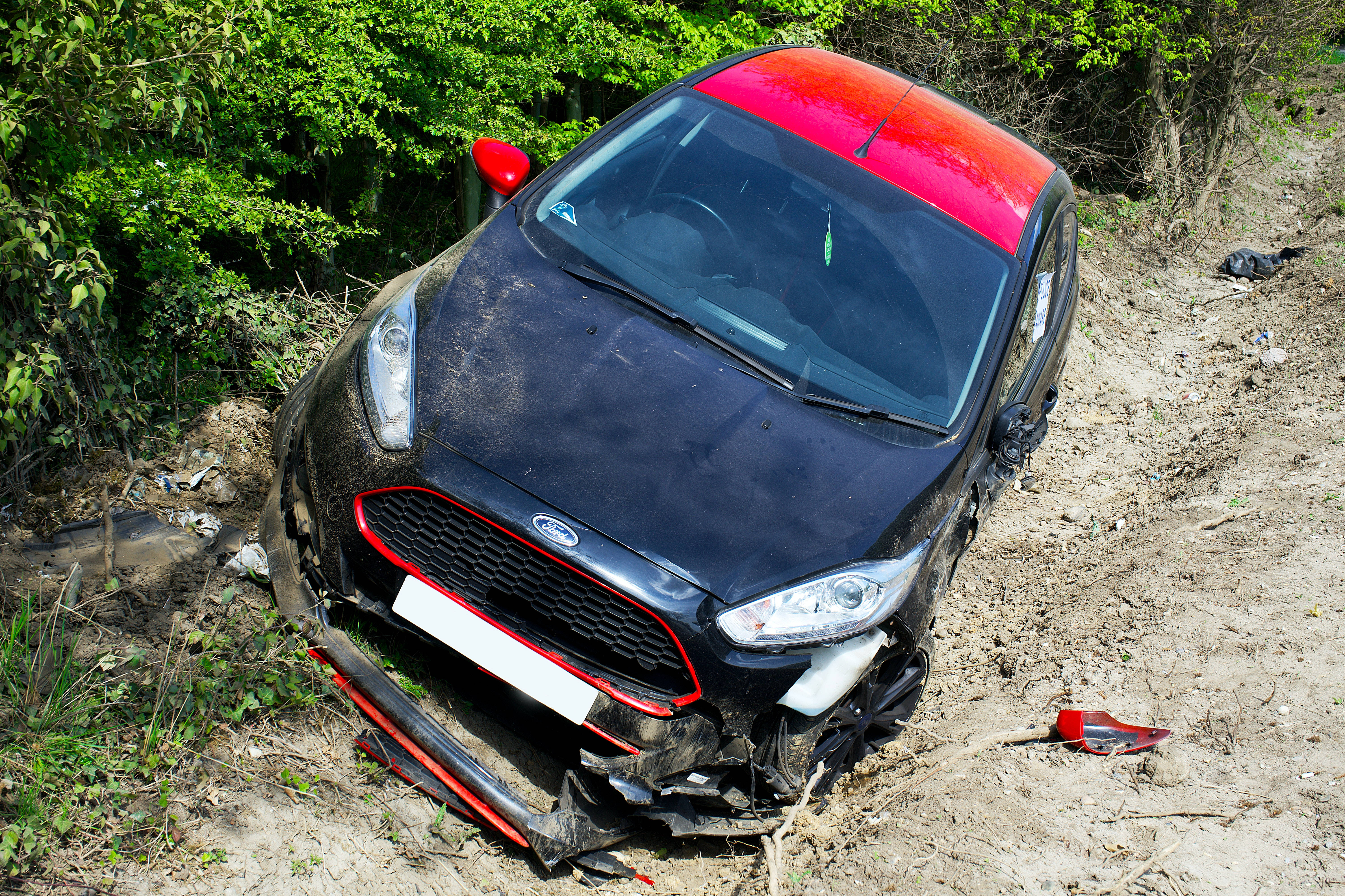car crash 桌布 壁紙 — 一輛黑色和紅色的福特汽車在泥濘的地形中損毀，周圍環繞著綠色樹葉，在明亮的日光下。