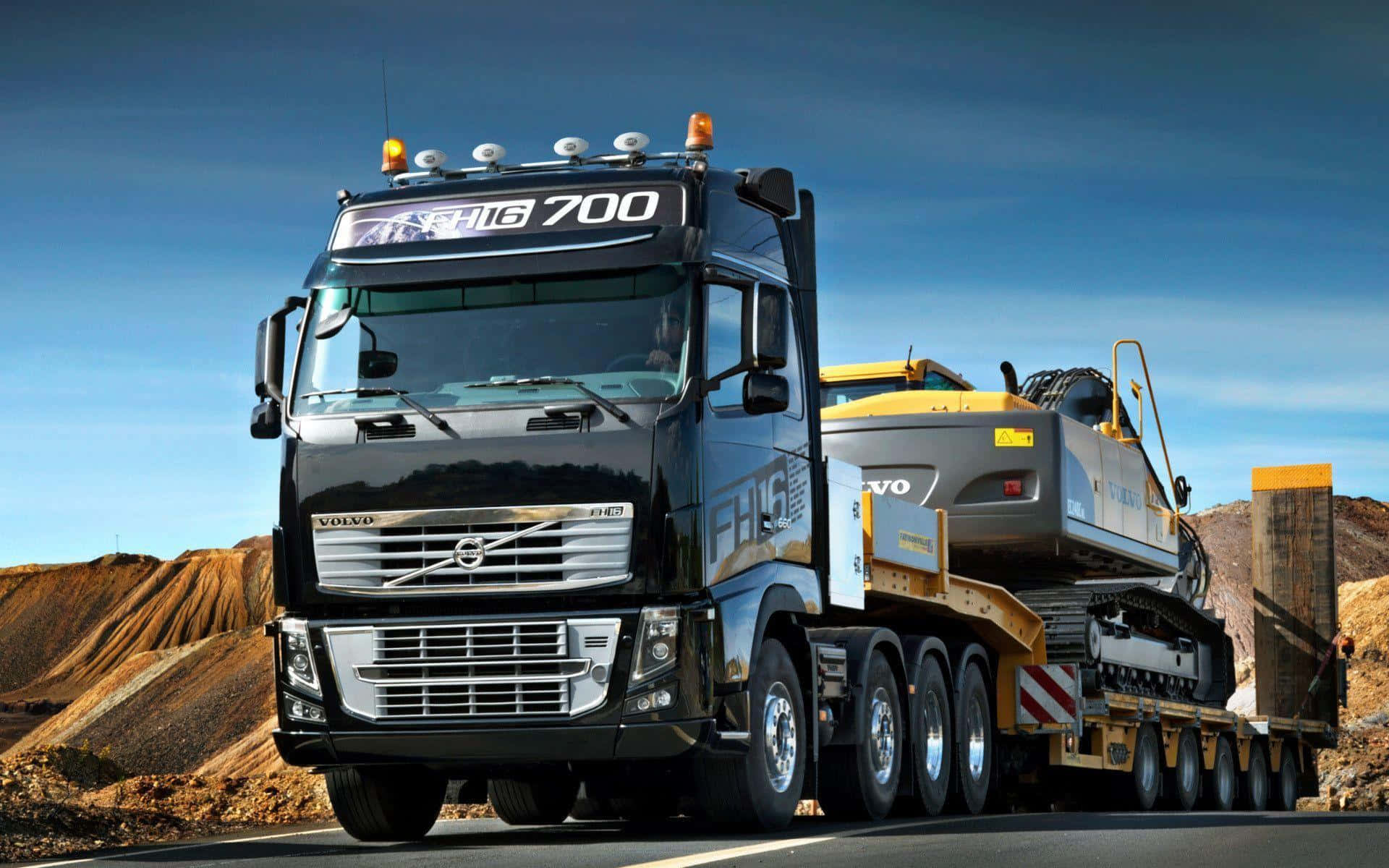 Big Truck 桌布 壁紙 — 一輛黑色富豪FH16 700重型貨車在晴朗藍天下拖著一台黃色挖土機穿越沙漠般的地形。