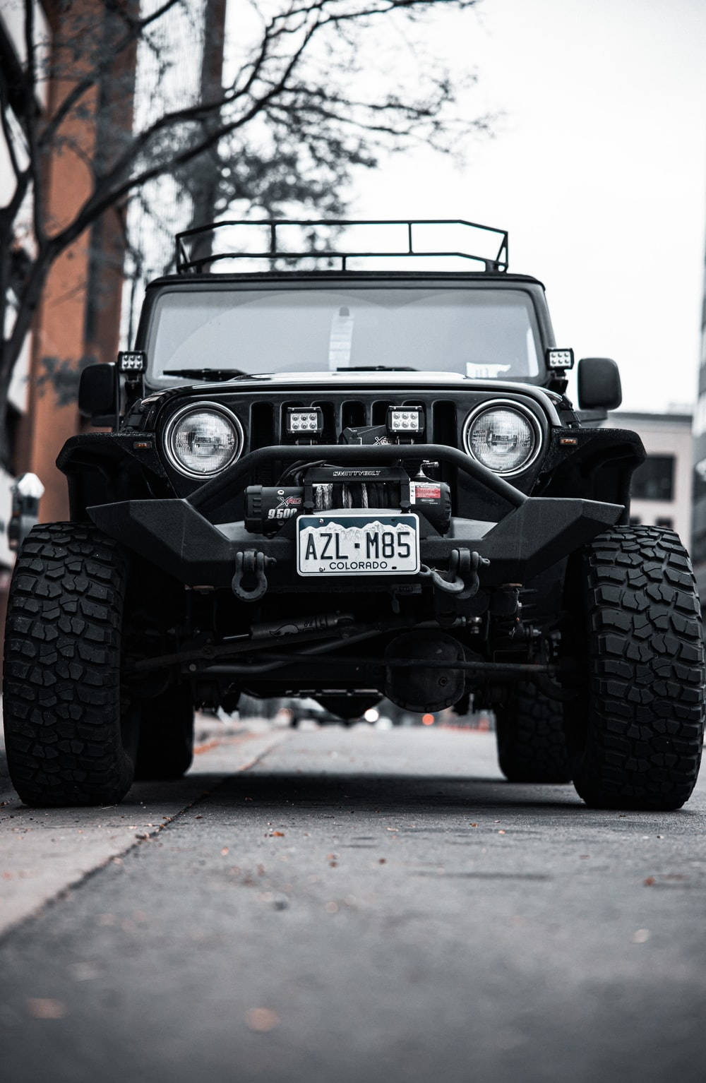 Wrangler 桌布 壁紙 — 一輛黑色改裝Jeep Wrangler，配備大型輪胎和車頂行李架，停泊在陰天城市街道上。