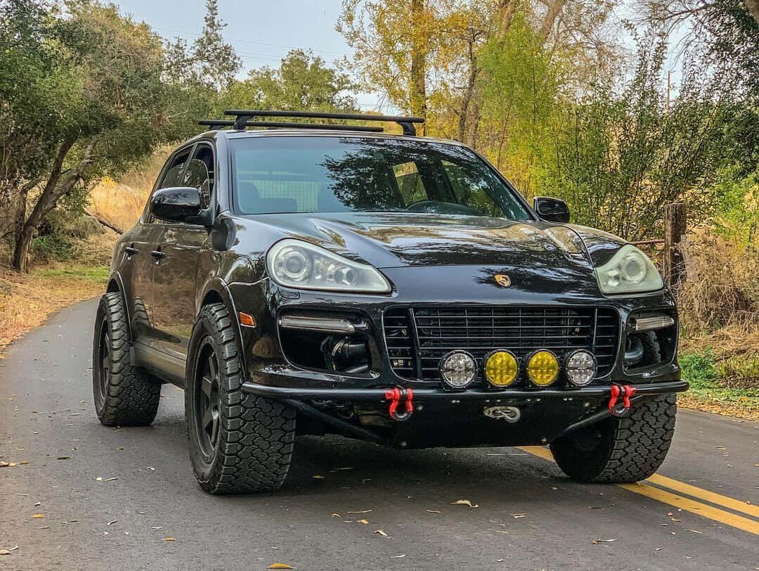 Off-road Car 桌布 壁紙 — 一輛黑色改裝保時捷卡宴SUV，配備越野輪胎和黃色輔助燈，停泊在林蔭大道上。