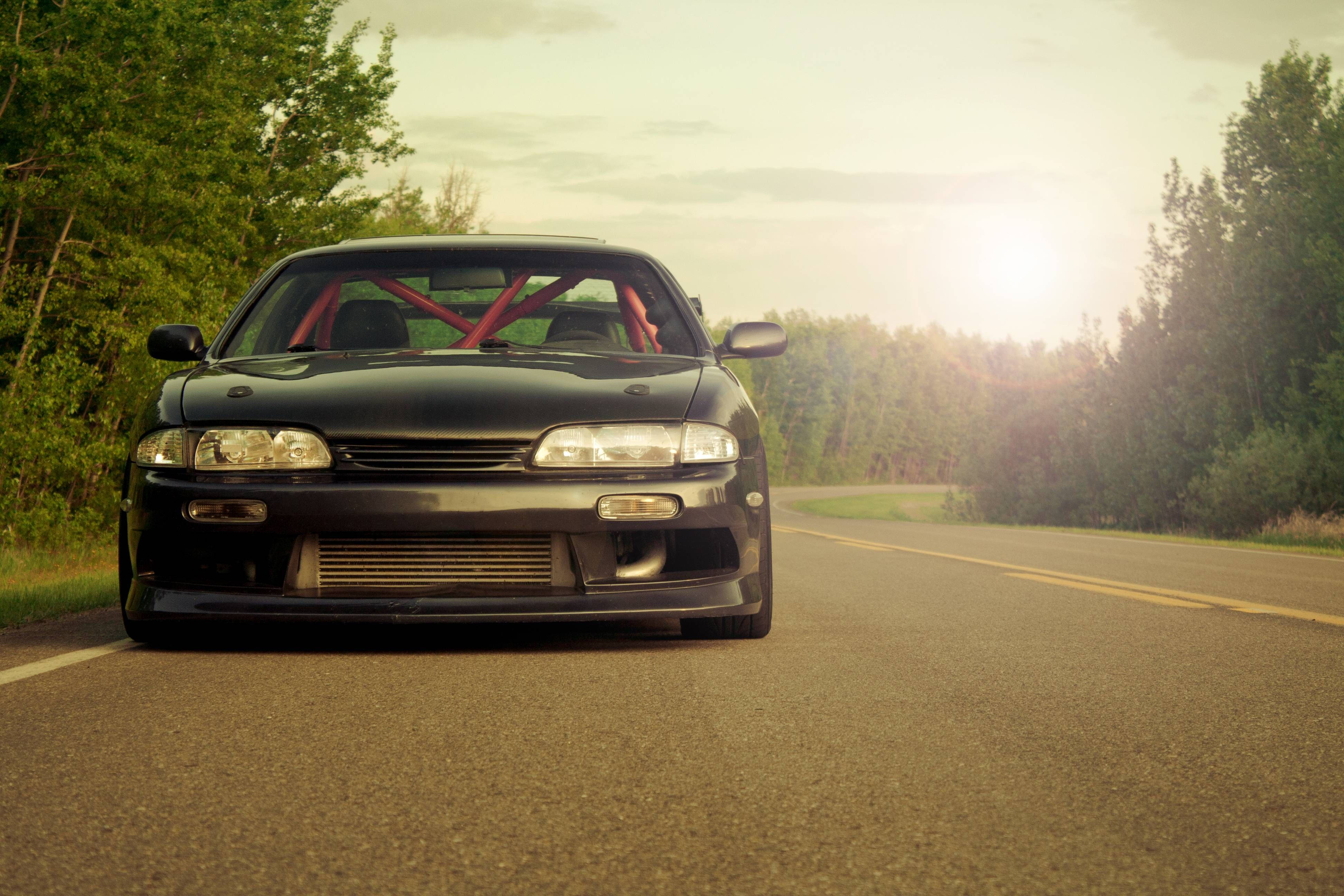 Nissan Silvia S14 桌布 壁紙 — 一輛黑色改裝跑車配備紅色防滾架，停在沐浴在金色日落光線中的森林道路上。