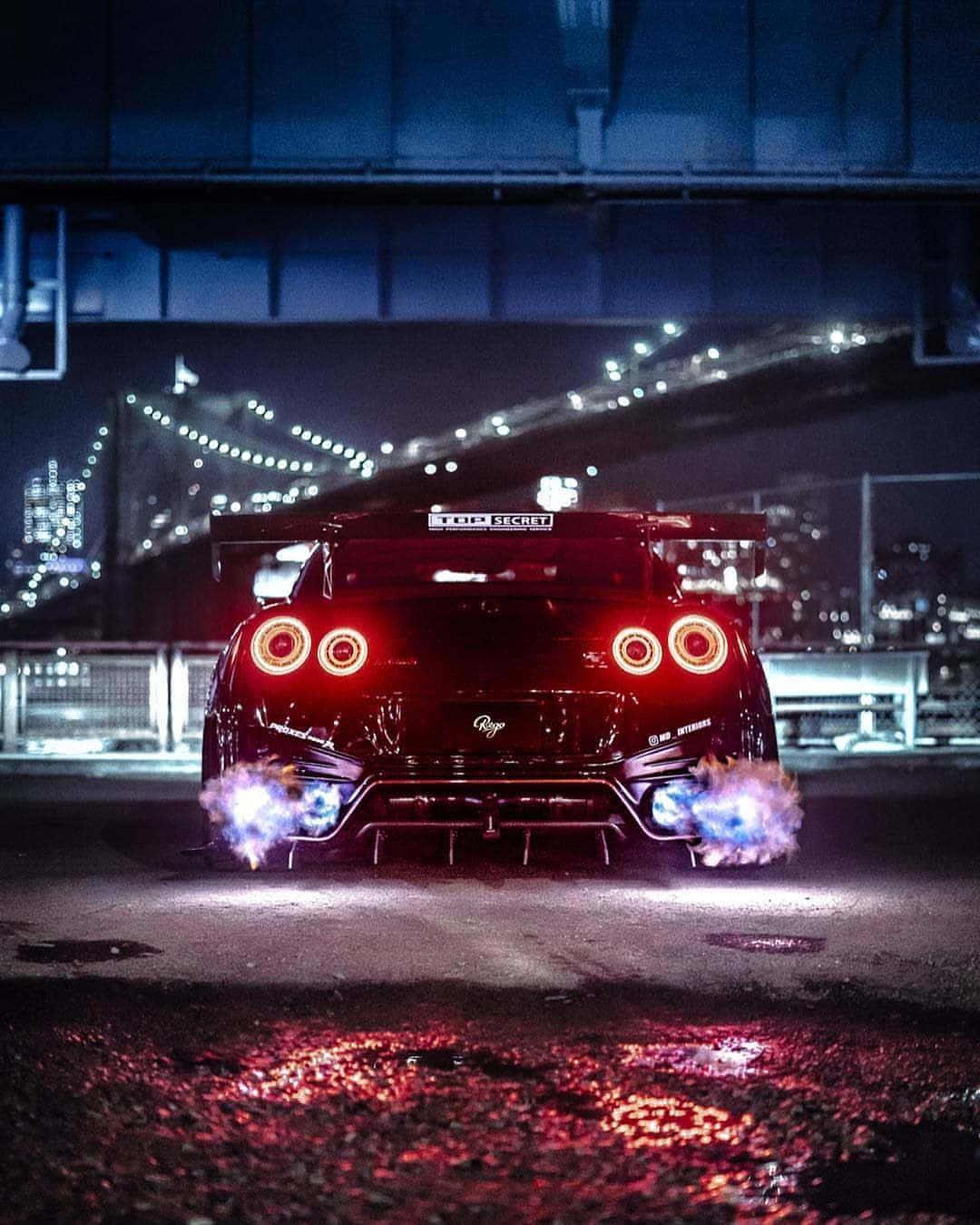 Nissan 桌布 壁紙 — 一輛黑色日產GT-R，帶有發光的紅色尾燈和藍色火焰排氣管，在夜間於城市橋下行駛。