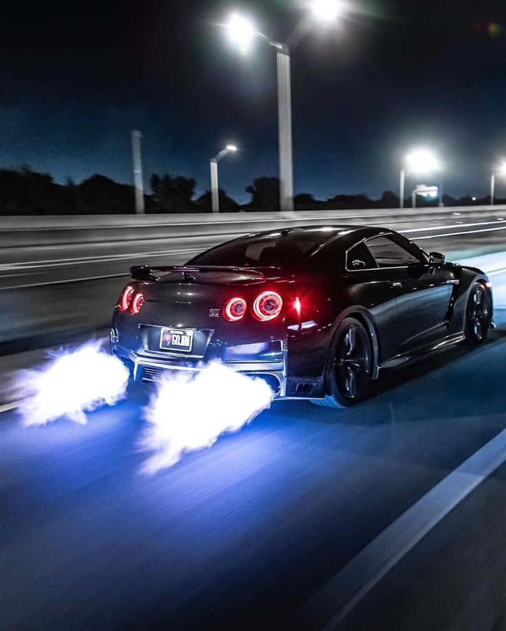 日產 桌布 壁紙 — 一輛黑色日產GT-R在夜間於照明高速公路上甩尾，後輪冒出發光的藍色煙霧。