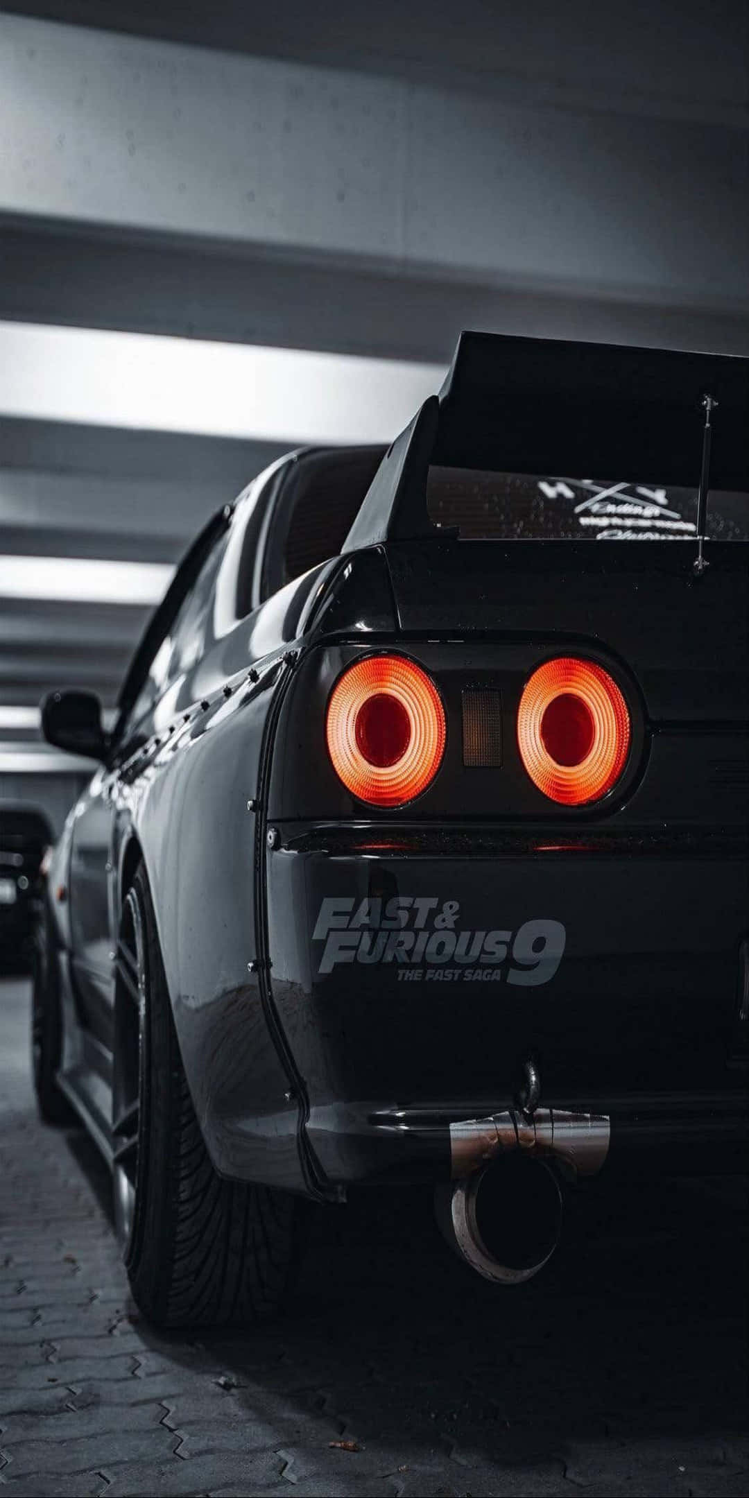 Pixel 3 Fast And Furious 桌布 壁紙 — 一輛黑色日產Skyline R34，帶有發光的橙色尾燈，停在光線昏暗的地下車庫中。