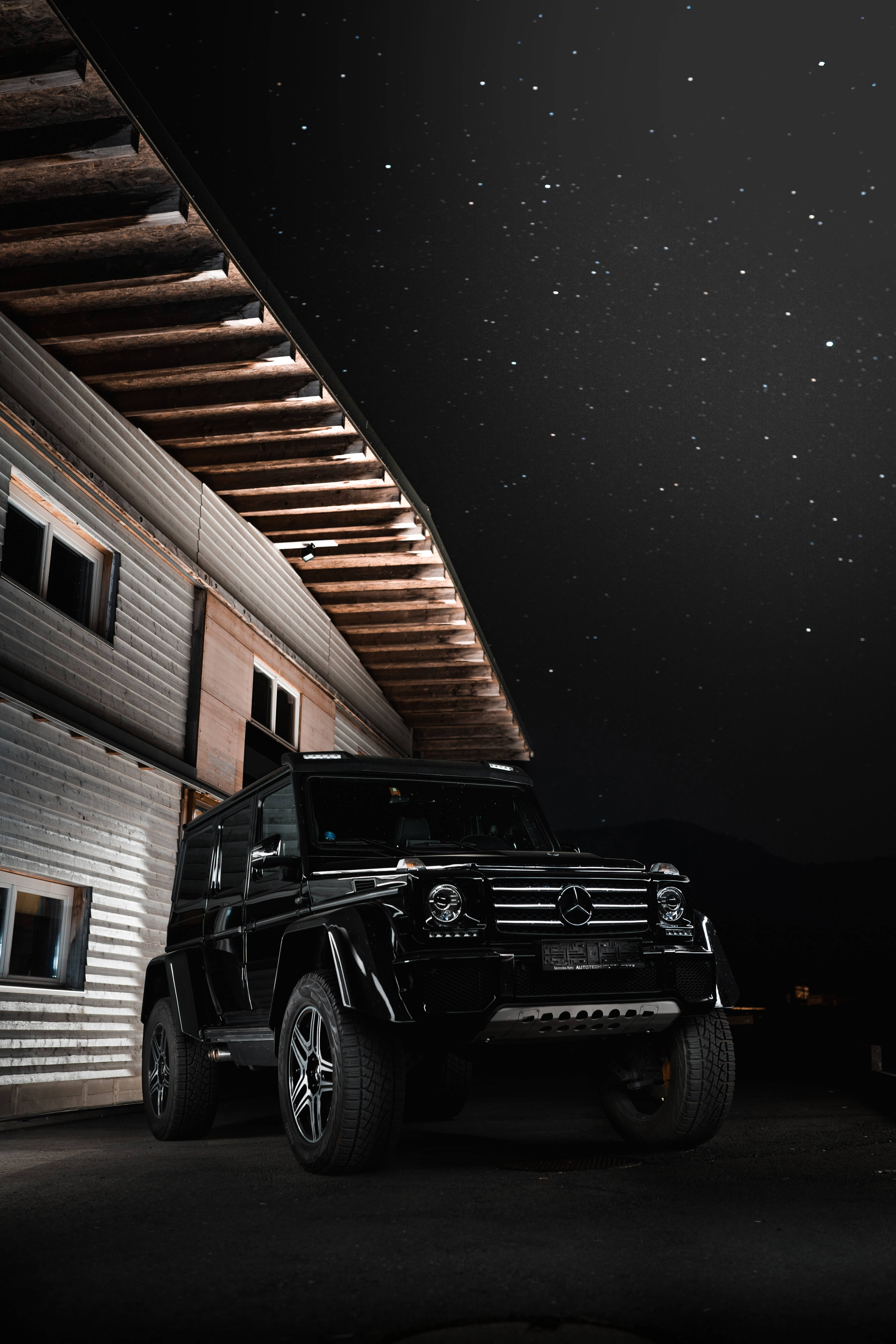 跑車 桌布 壁紙 — 一輛黑色梅賽德斯G-Wagen停泊在星空夜空下，旁邊是一座帶有照明木樑的現代建築。