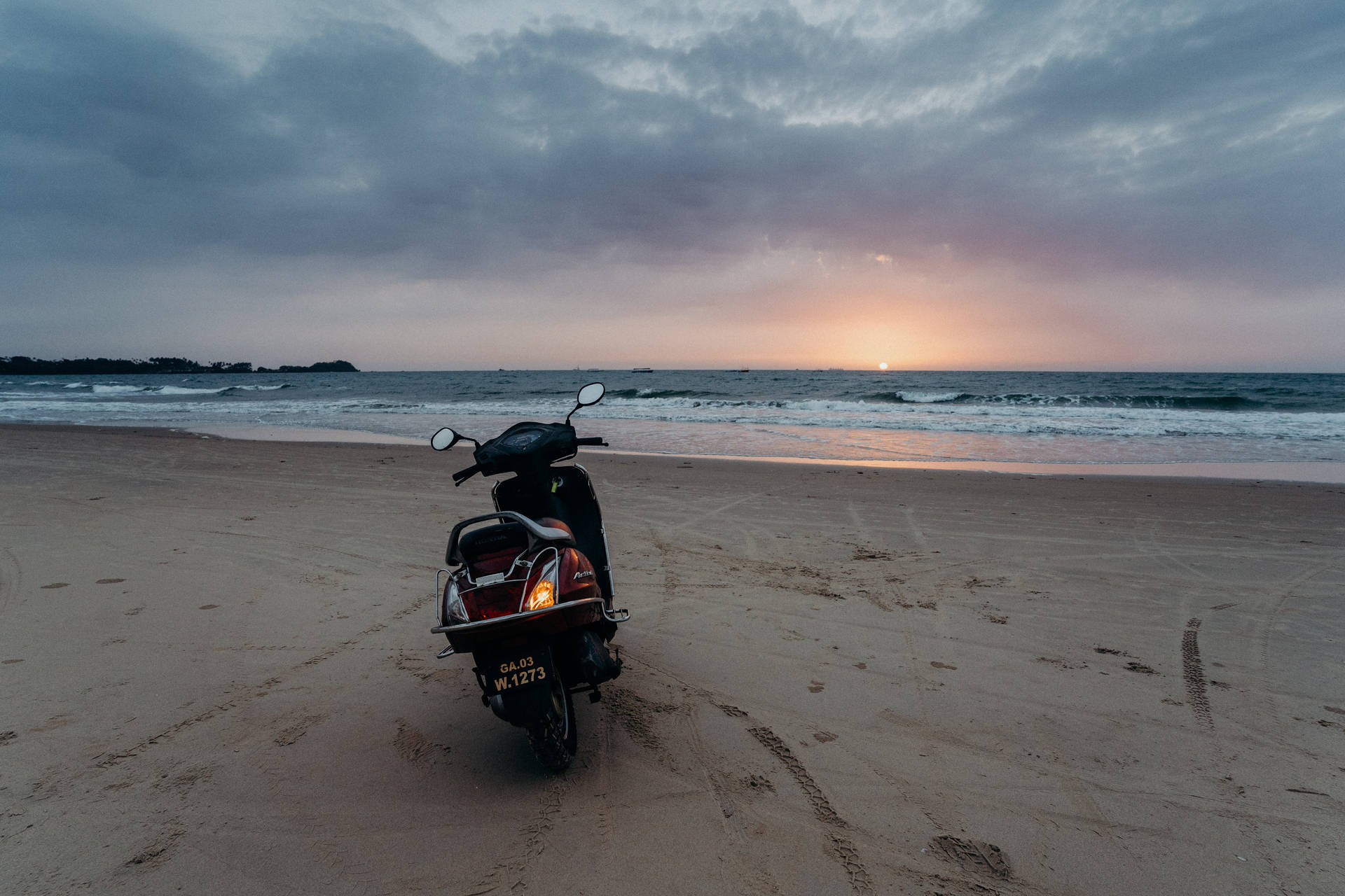  壁紙 — 一輛黑色機車停放在沙灘上，夕陽西下，海浪拍打，多雲的天空閃耀著橙色和紫色。