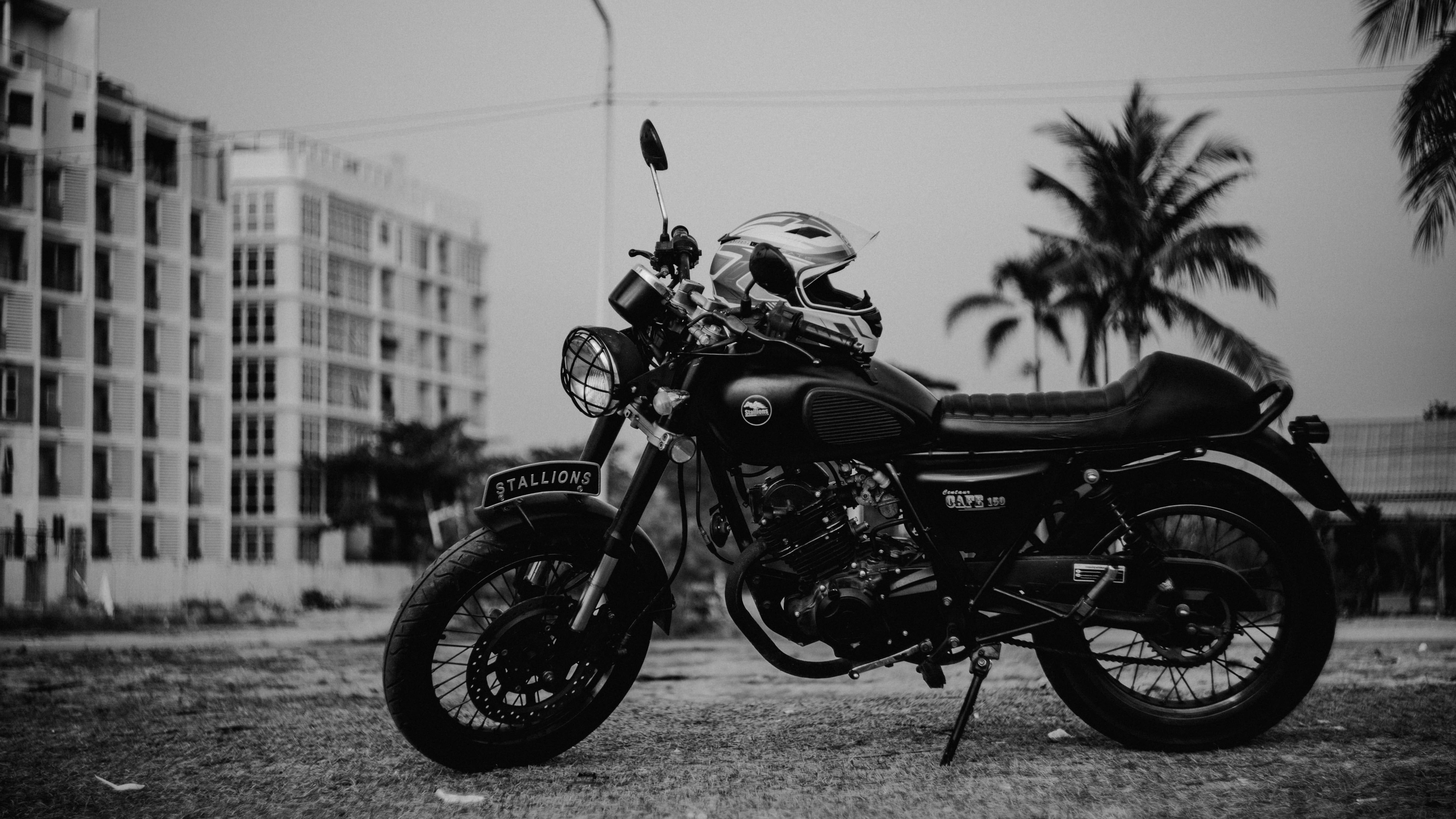 機車 桌布 壁紙 — 一輛黑色機車連同安全帽停放在側腳架上，背景是單色城市景觀，有棕櫚樹和建築物。