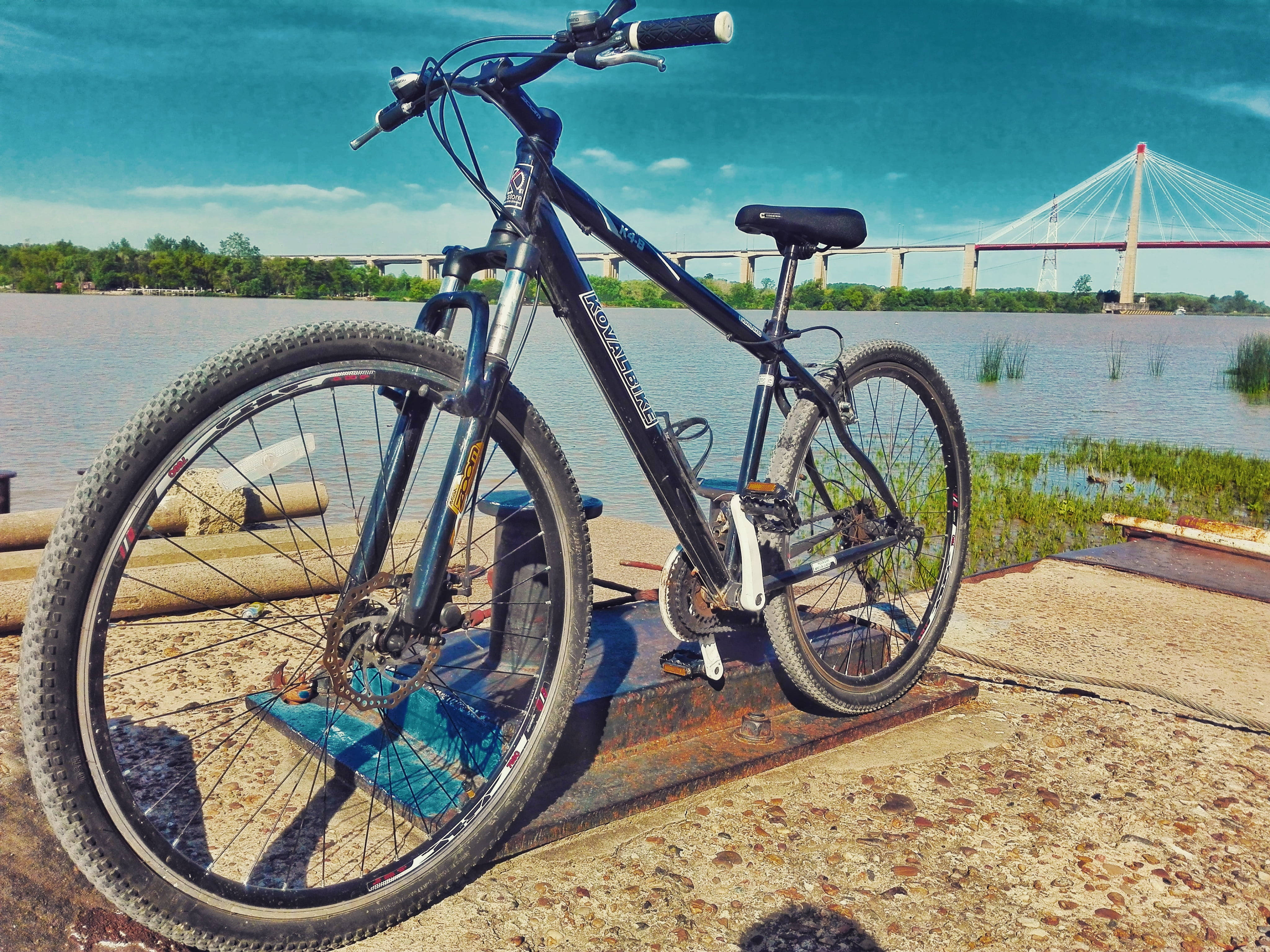 Mountain Bike 桌布 壁紙 — 一輛黑色登山車停放在生鏽的金屬平台上，旁邊是一條平靜的河流，一座現代化的纜索橋在明亮的藍天下橫跨水面。