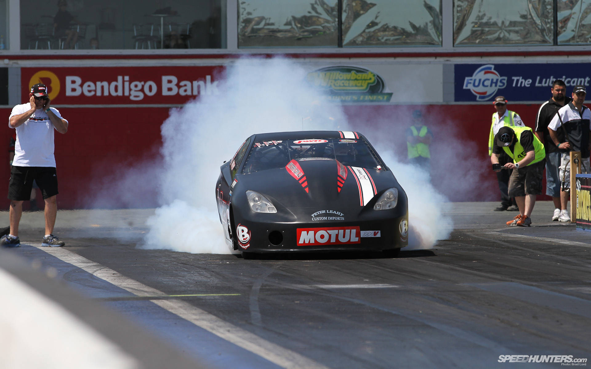 Racing 桌布 壁紙 — 一輛黑色直線加速賽車在柏油賽道上進行燒胎表演，揚起濃厚白煙，背景有觀眾和品牌橫幅。
