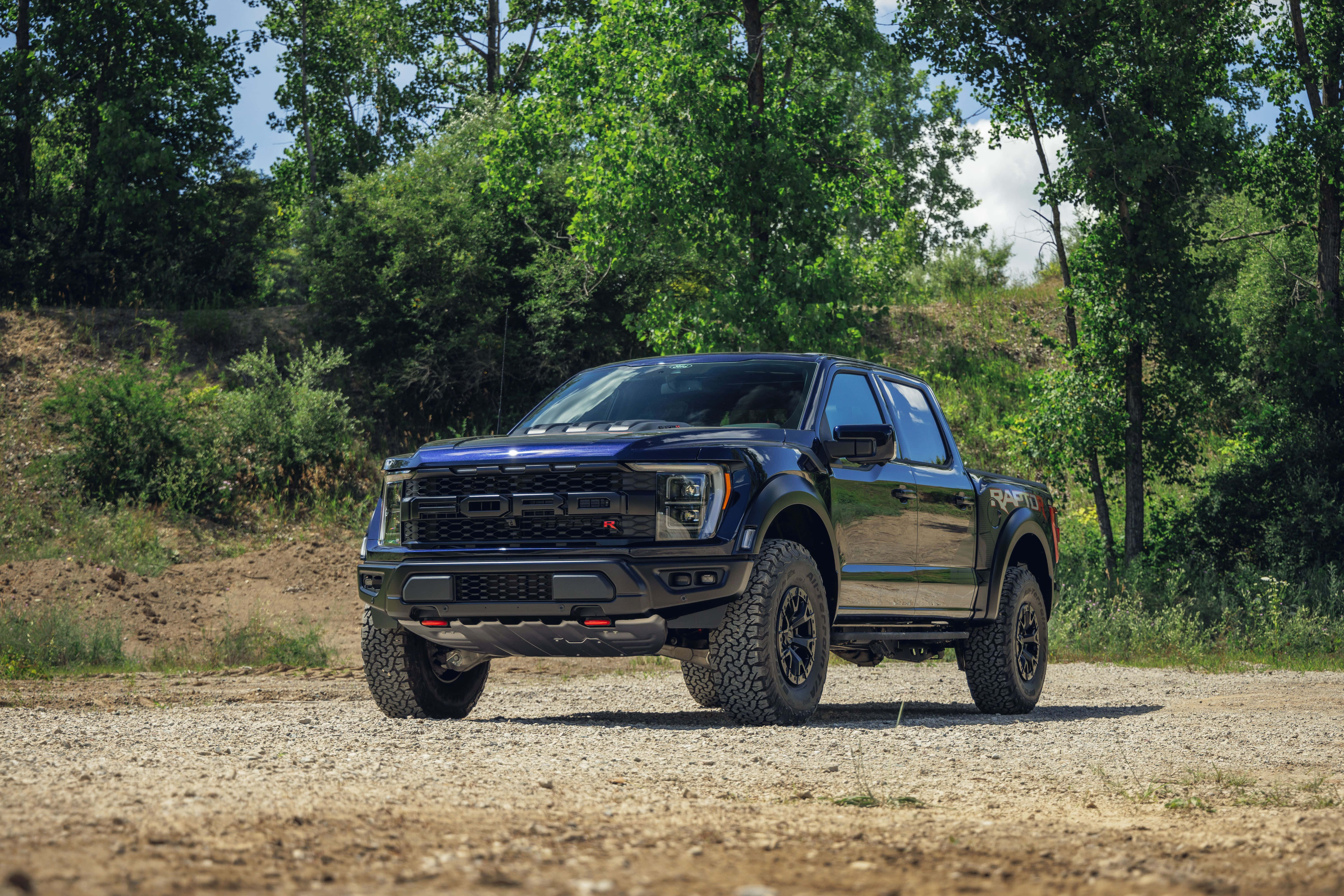 森林 桌布 壁紙 — 一輛黑色福特 F-150 Raptor 貨車大膽地停在碎石路上，周圍是茂密的綠樹，頭頂是明亮的藍天。