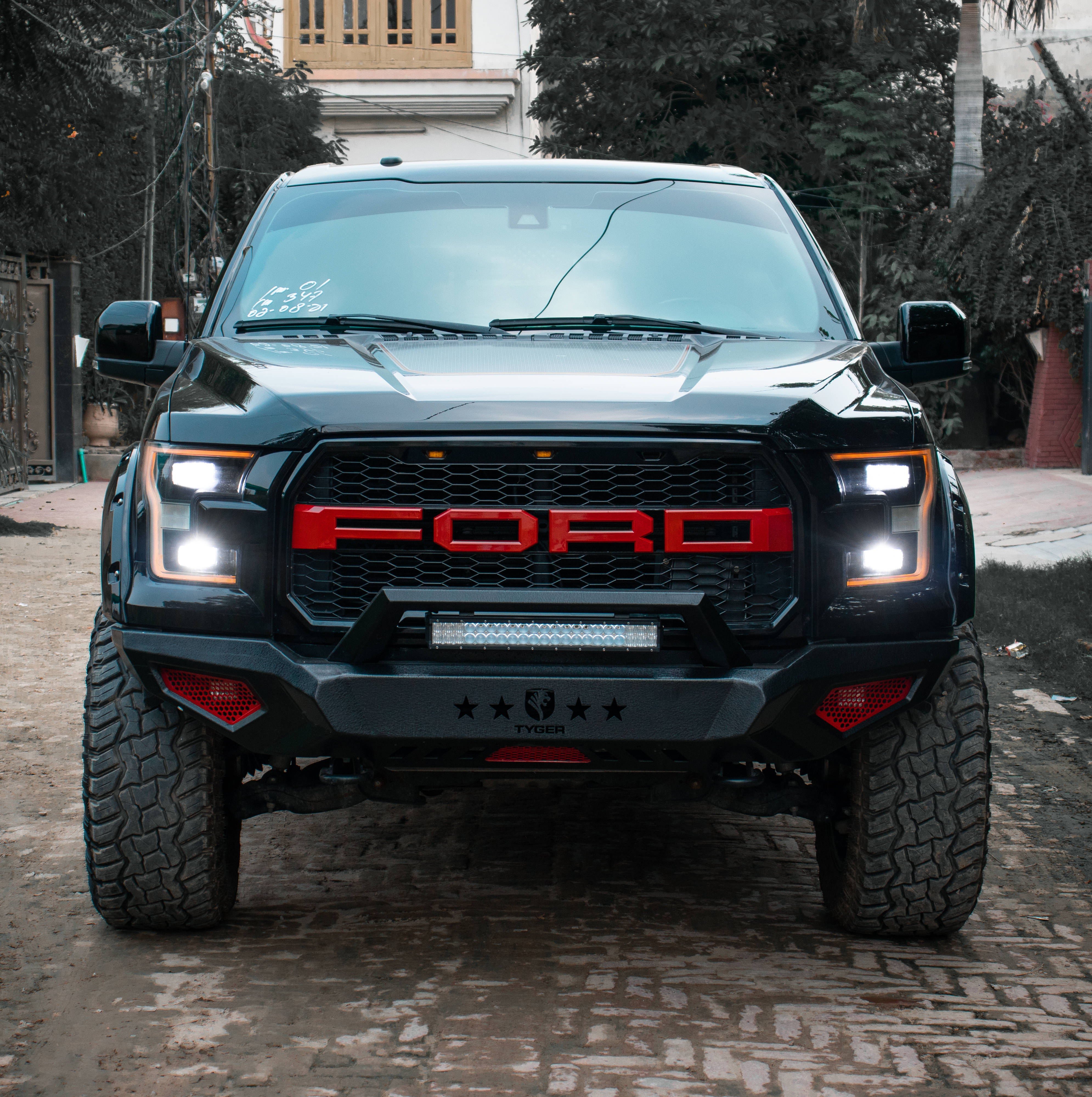 Raptor 桌布 壁紙 — 一輛黑色福特F-150 Raptor帶有紅色點綴和發光車頭燈，停在鵝卵石街道上，周圍環繞著樹木和建築物。