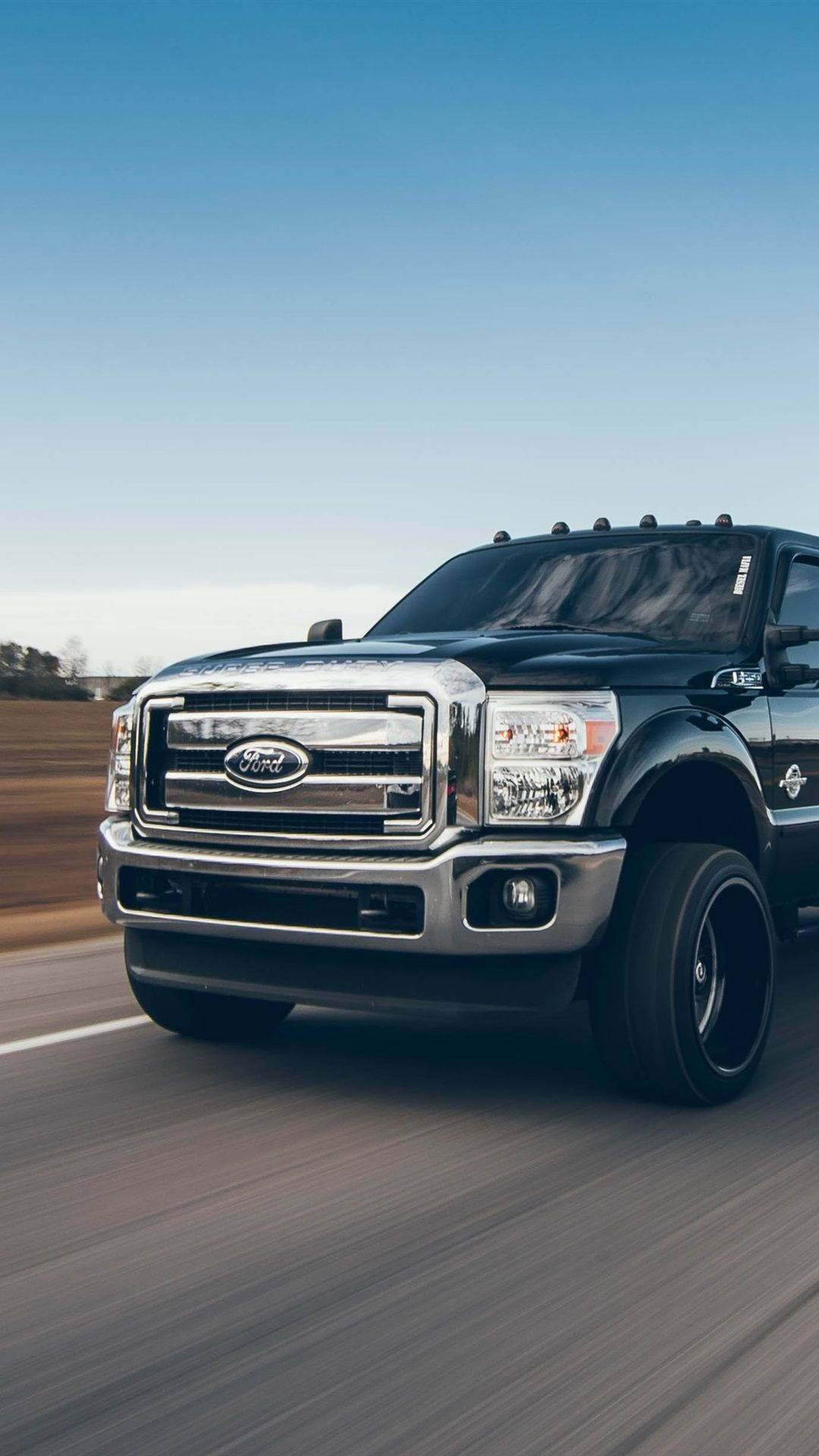 ford iPhone 桌布 壁紙 — 一輛黑色福特F-150卡車在晴朗藍天下於鄉間道路高速行駛，柏油路面呈現動態模糊效果。