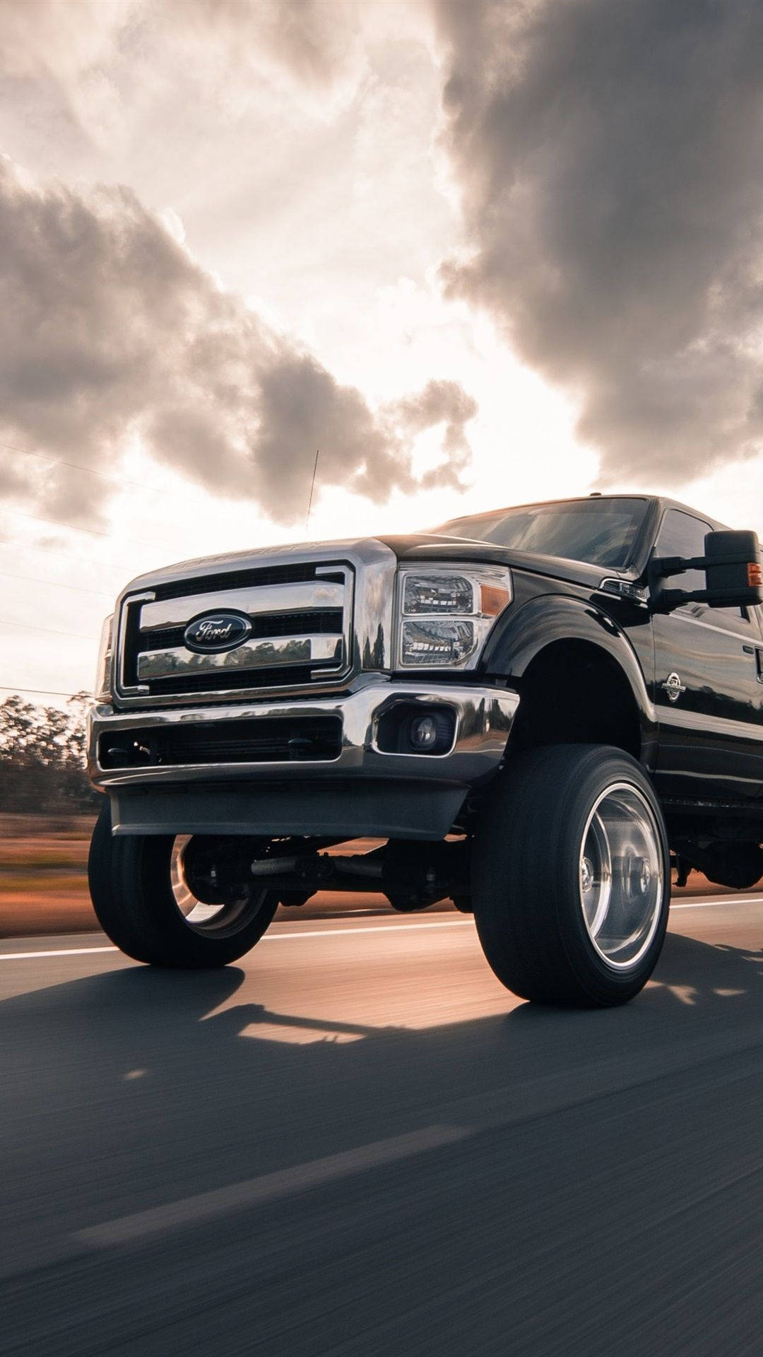 ford iPhone 桌布 壁紙 — 一輛黑色福特F-150皮卡車在日落時分戲劇性的多雲天空下在高速公路上疾馳。