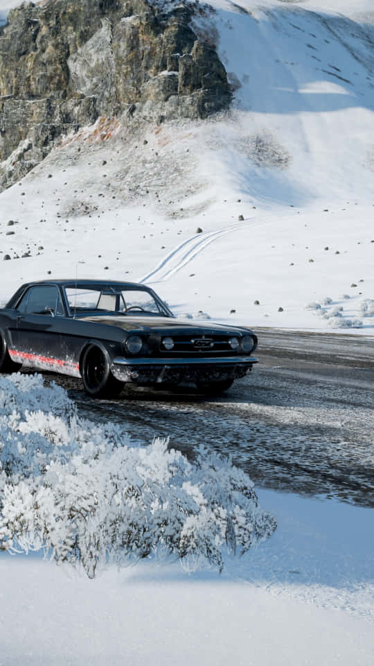 Android Forza Horizon 4 桌布 壁紙 — 一輛黑色經典肌肉車在白雪皚皚的山路上行駛，兩旁是結霜的灌木叢和岩石峭壁，在明亮的日光下。