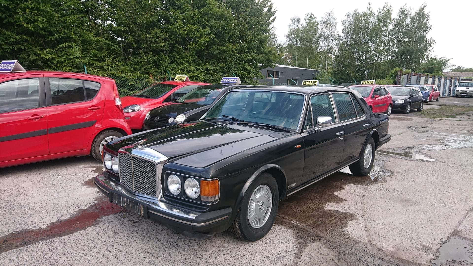 Bentley Eight 桌布 壁紙 — 一輛黑色經典賓利轎車停在潮濕的停車場，周圍是色彩繽紛的計程車和綠樹。