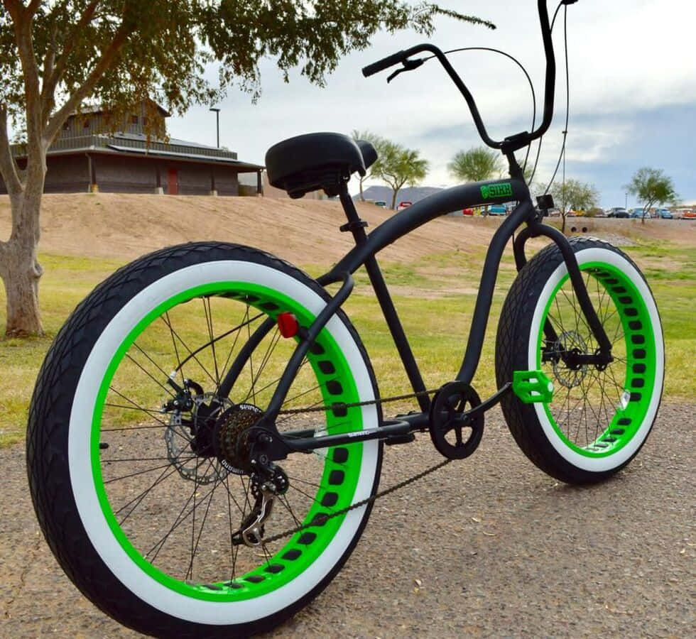 Beach Cruiser 桌布 壁紙 — 一輛黑色胖胎車，帶有螢光綠色輪圈和白色輪胎，停在碎石地上，靠近一棵樹和木建築。