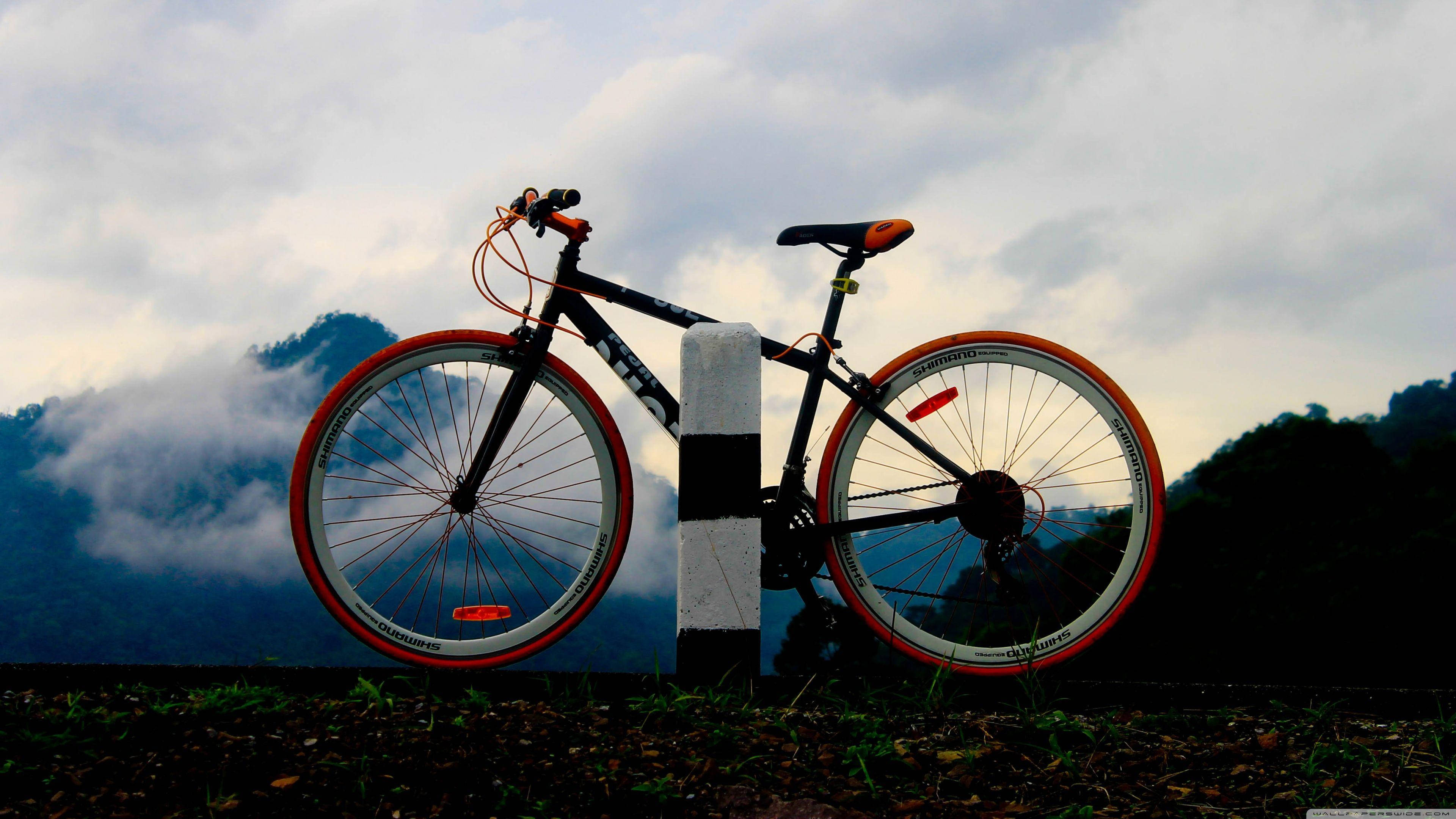 4K bike 桌布 壁紙 — 一輛黑色自行車配橙色輪圈，靠在一根條紋柱上，背景是多雲的山景。