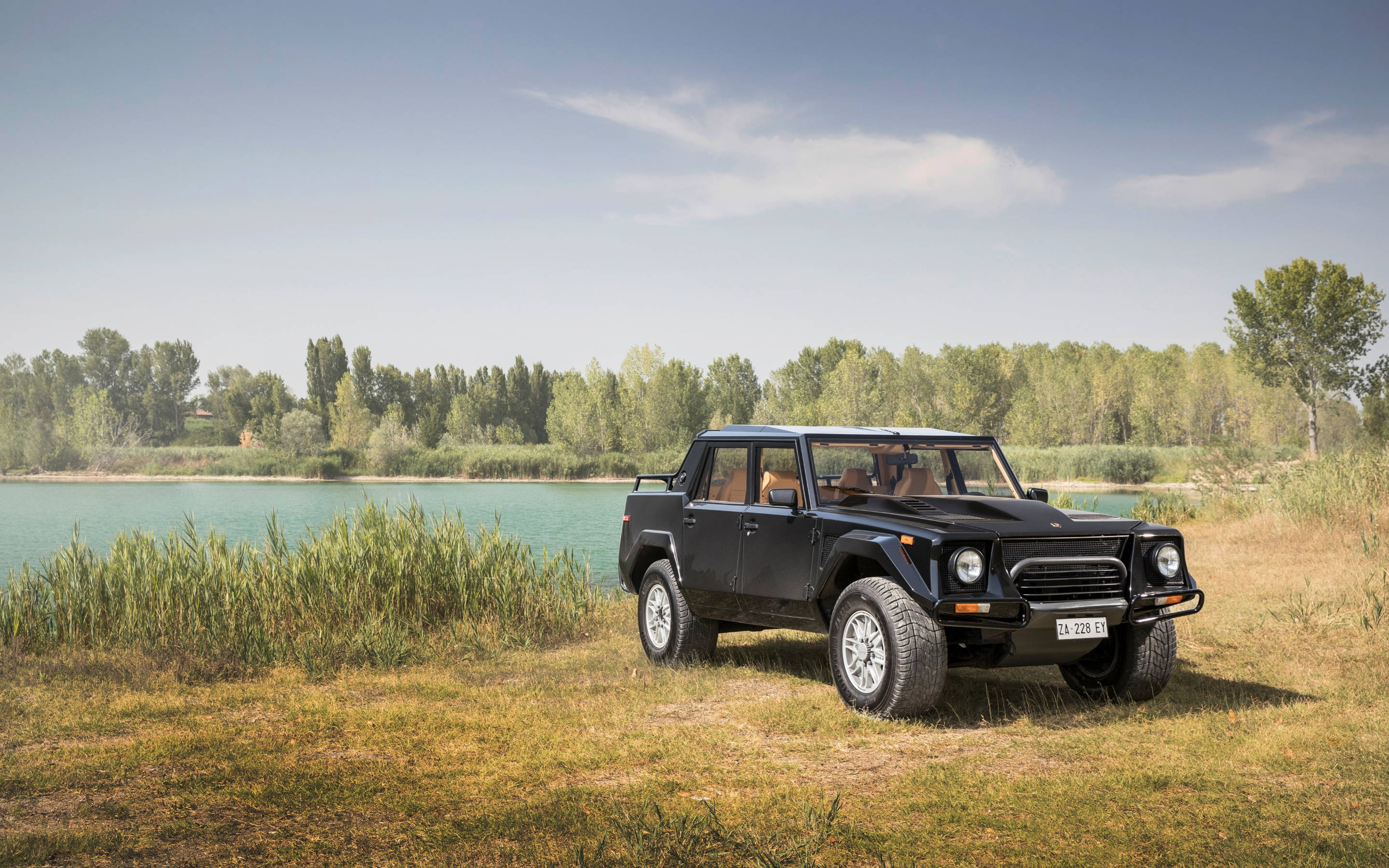 lambo 桌布 壁紙 — 一輛黑色藍寶堅尼 LM002 SUV 停泊在平靜的土耳其藍色湖泊旁的草地上，背景有樹木，天空清澈湛藍。