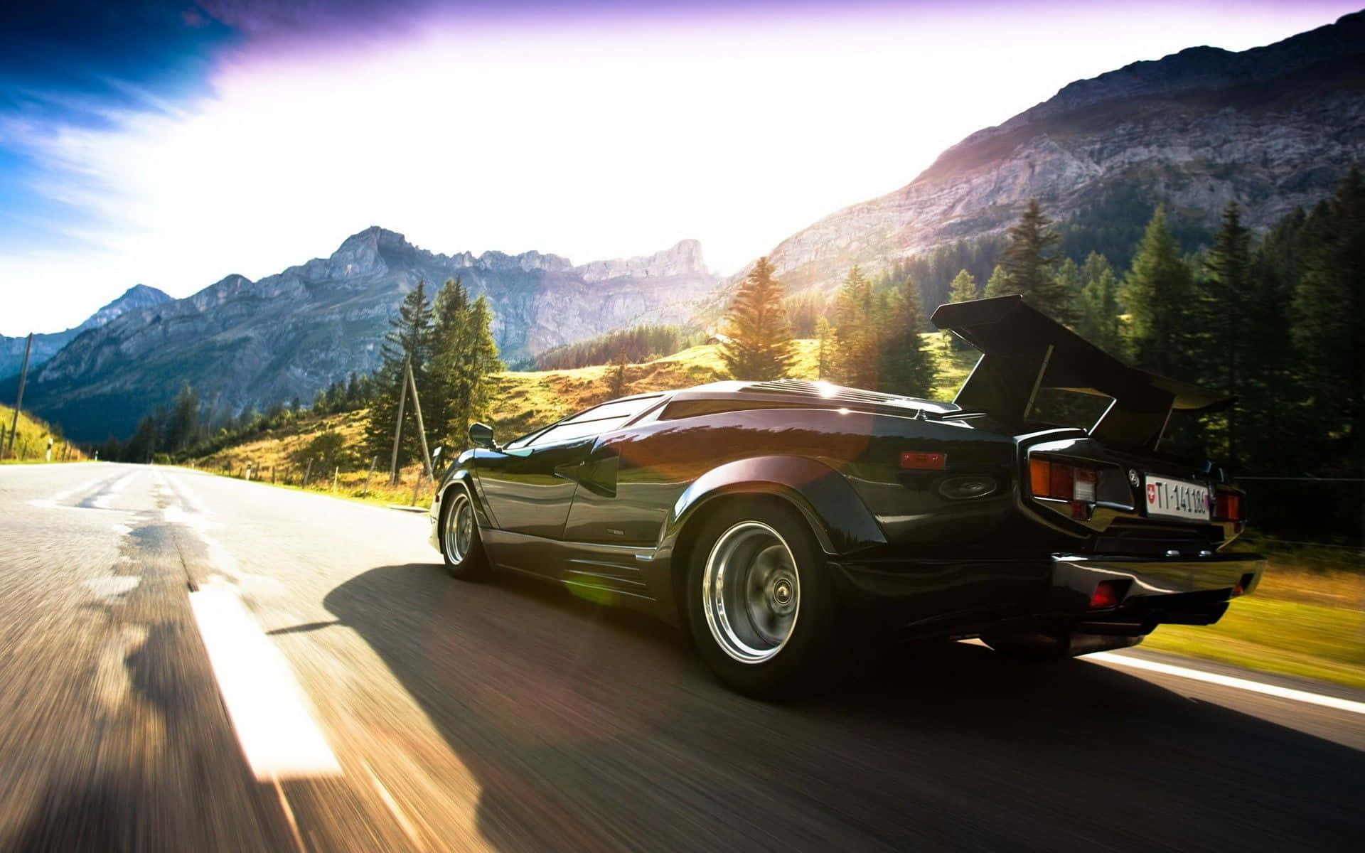 Lamborghini Countach 桌布 壁紙 — 一輛黑色藍寶堅尼Countach在蜿蜒的山路上疾馳，尾翼升起，陽光照射在崎嶇的山峰和松樹林上。