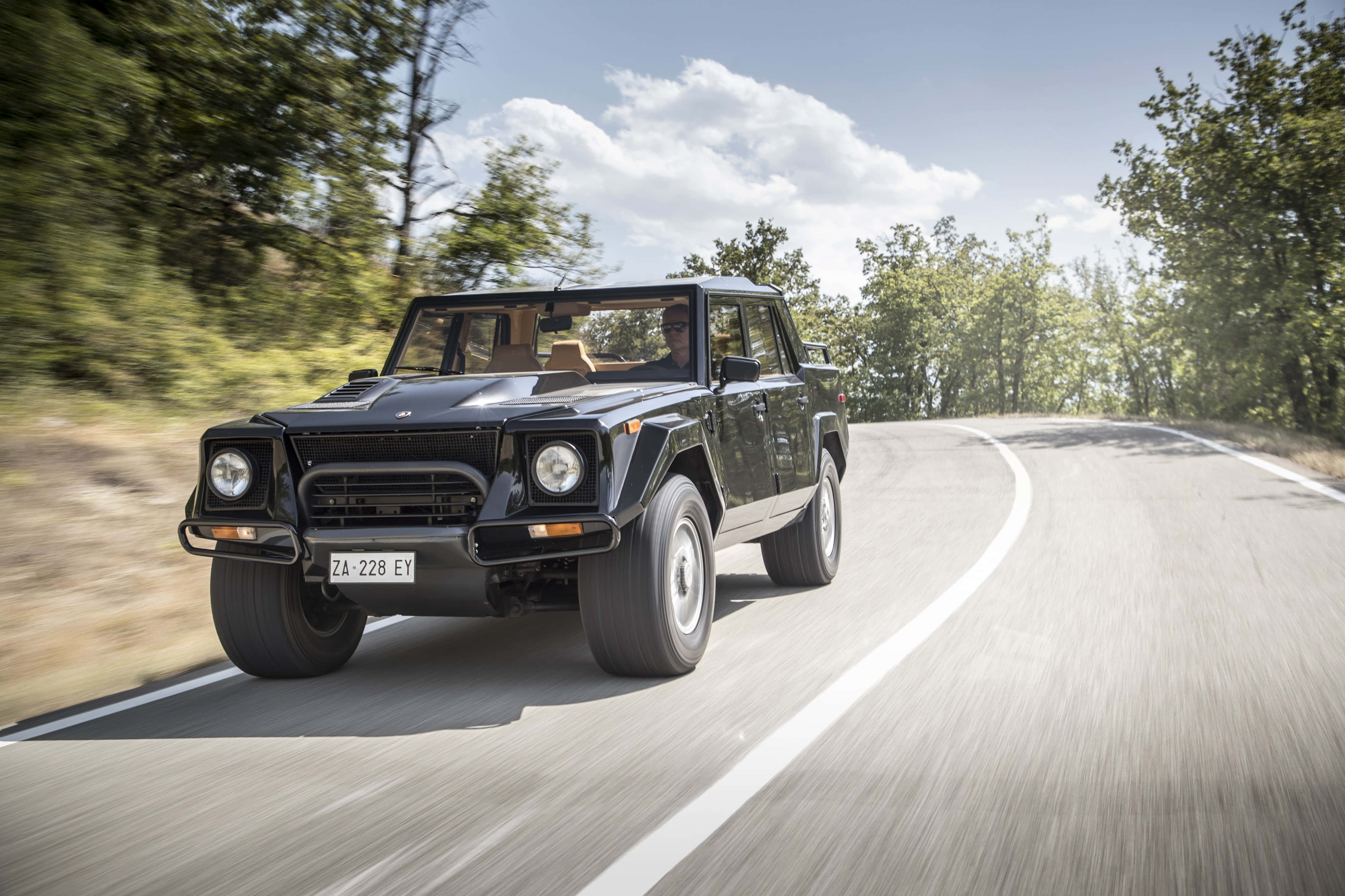 Lamborghini Lm002 桌布 壁紙 — 一輛黑色藍寶堅尼LM002 SUV在晴朗藍天和蓬鬆白雲下，沿著蜿蜒的森林道路飛馳。