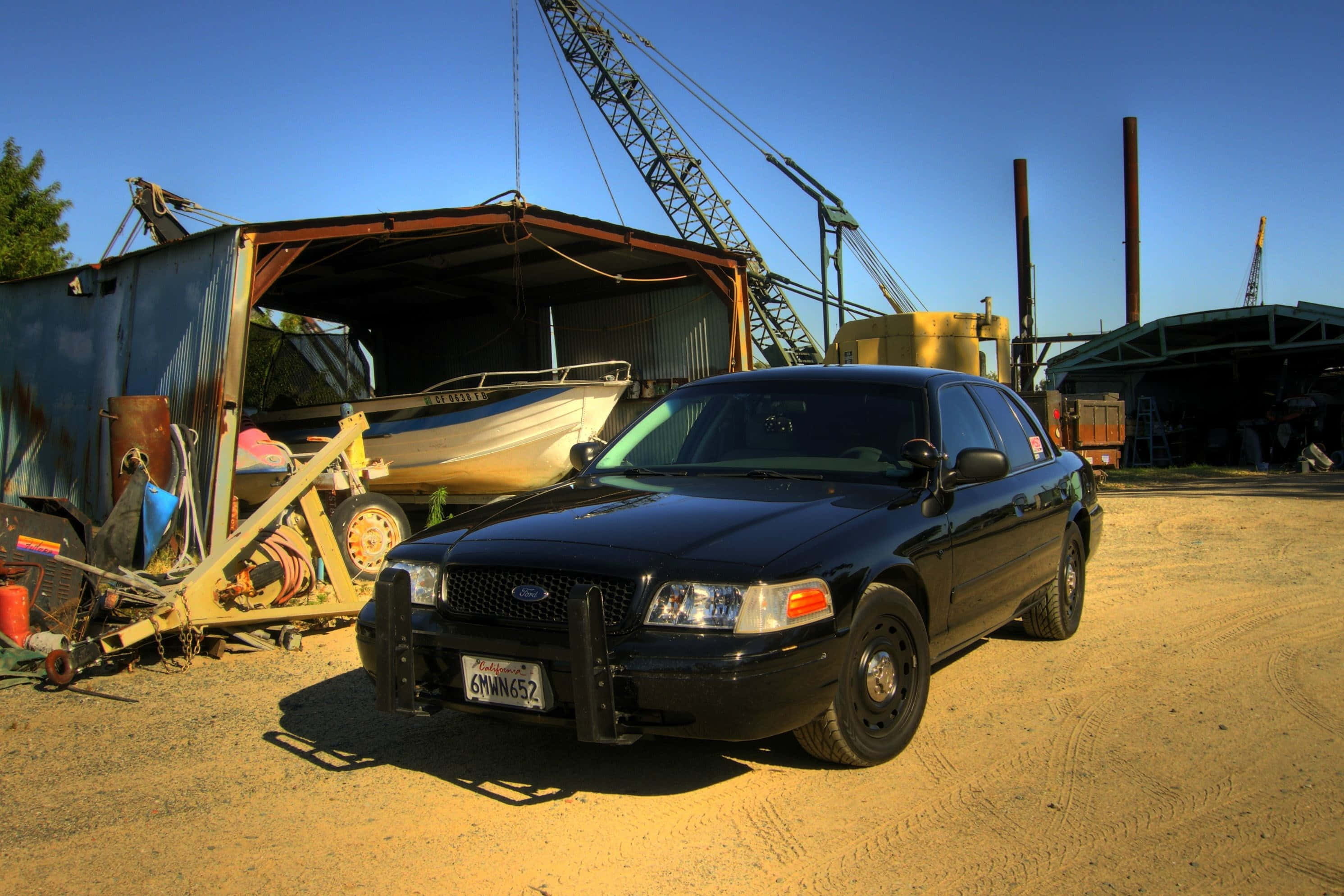 Ford Crown Victoria 桌布 壁紙 — 一輛黑色警車停泊在塵土飛揚的地面上，前面是生鏽的船和工業起重機，背景是晴朗的藍天。