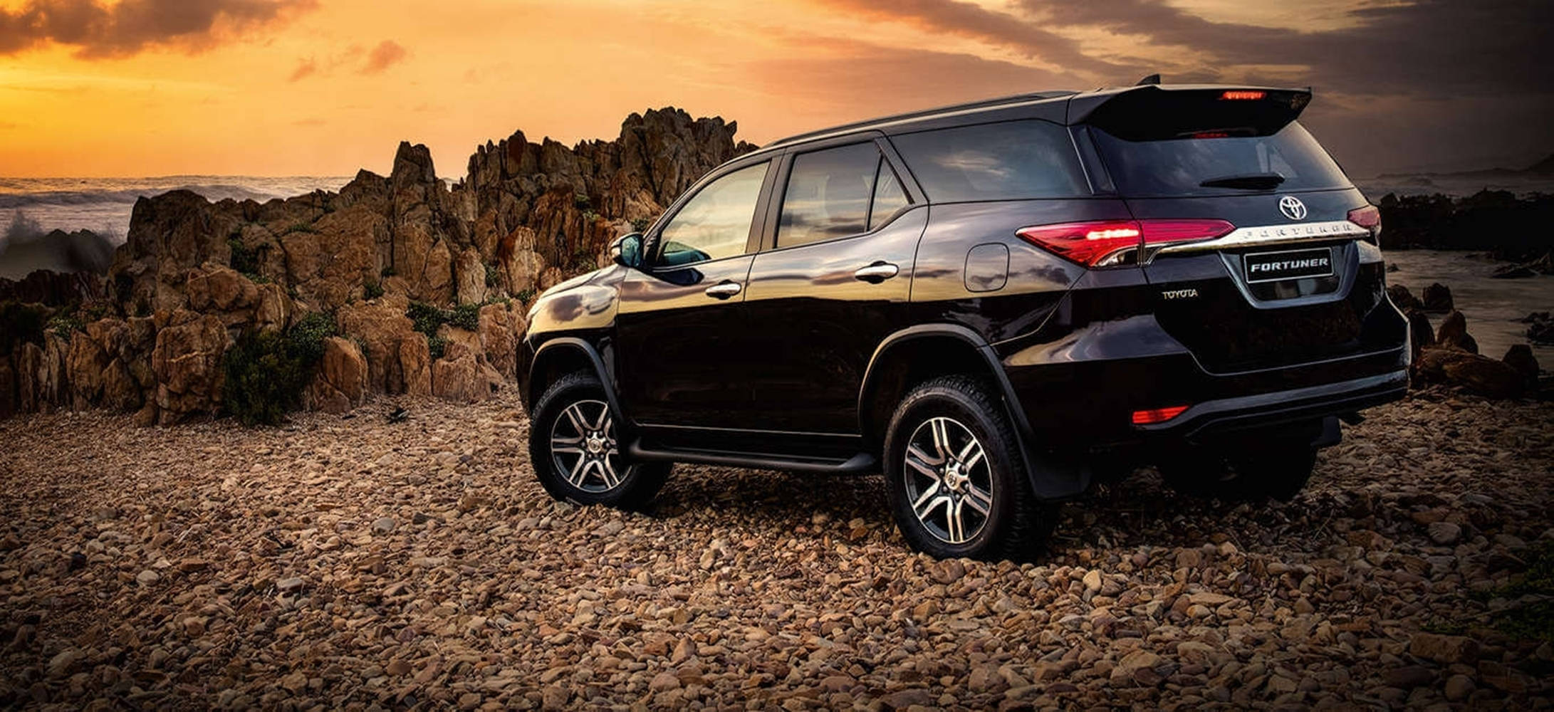 Toyota 桌布 壁紙 — 一輛黑色豐田 Fortuner SUV 停放在岩石地形上，背景是日落時分戲劇性的橙色天空和崎嶇的懸崖。