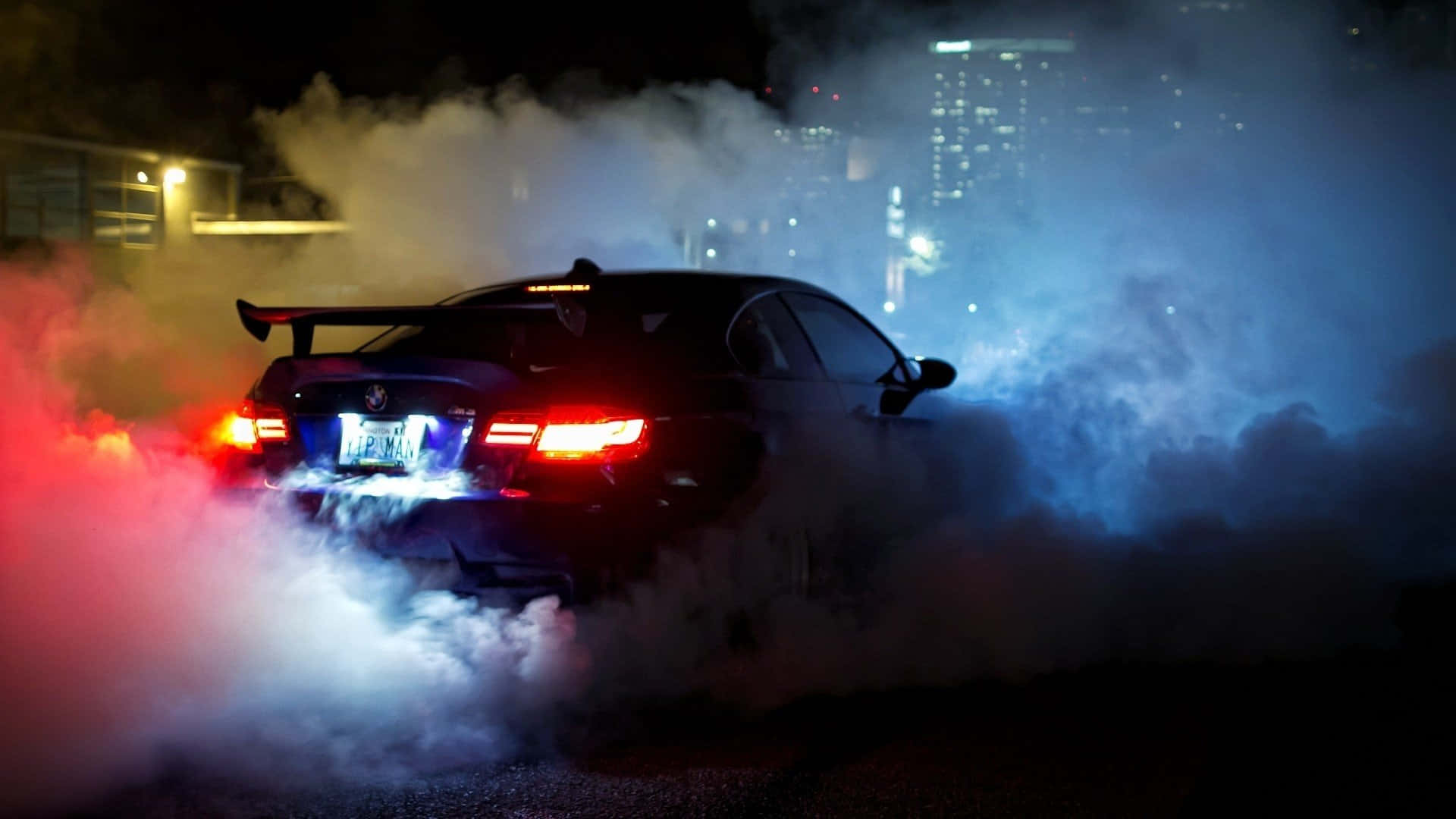 桌布 壁紙 — 一輛黑色跑車在夜間進行燒胎，揚起濃煙，被紅色和藍色燈光照亮，背景是城市天際線。