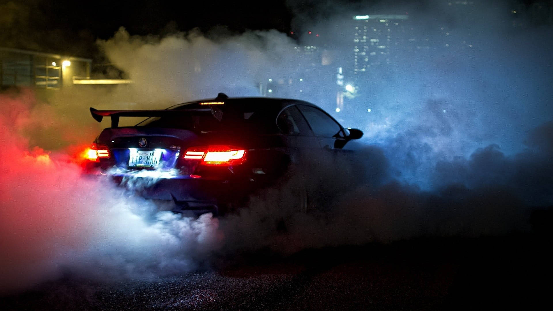 黑色跑車在夜間飄移，在城市中揚起紅色和藍色煙霧。 — BMW 動圖 壁紙