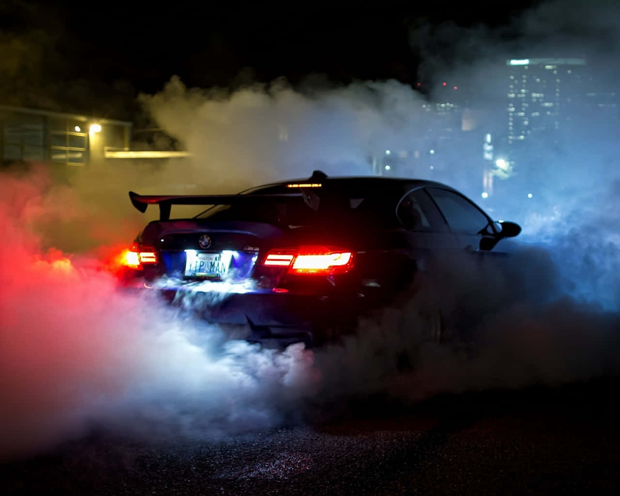 桌布 壁紙 — 一輛黑色跑車在夜間飄移，揚起被紅藍燈光照亮的煙霧，背景是城市天際線。