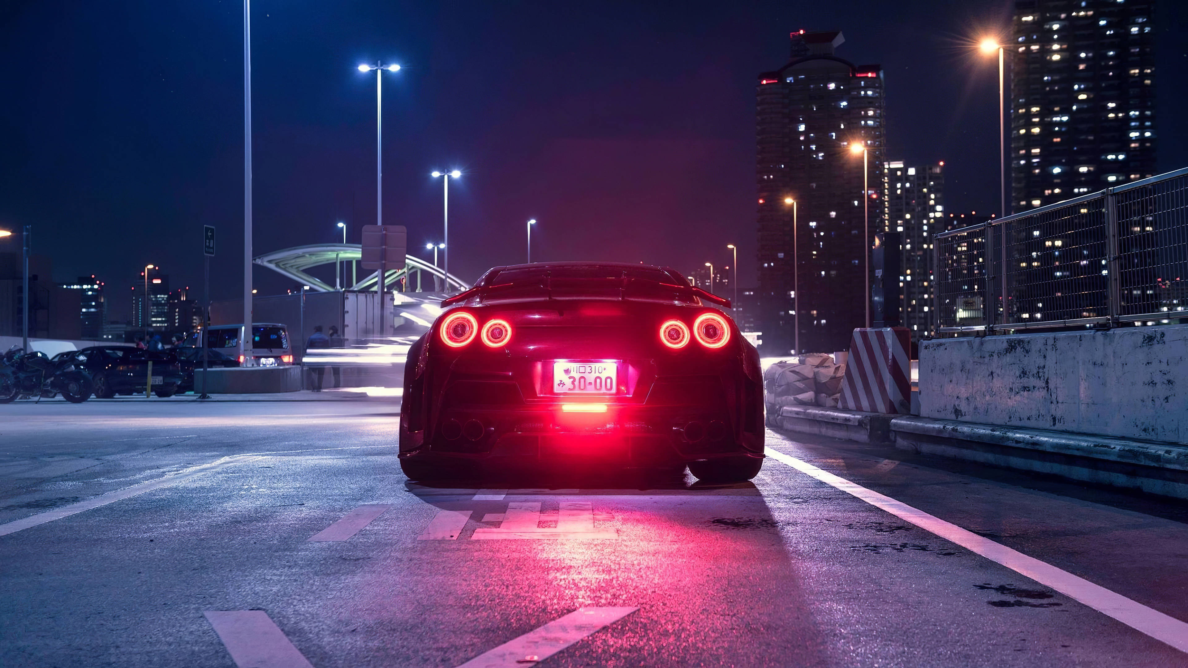 Nissan 桌布 壁紙 — 一輛黑色跑車帶有發光的紅色尾燈，夜晚停在濕漉漉的城市道路上，周圍是高聳的燈光摩天大樓。