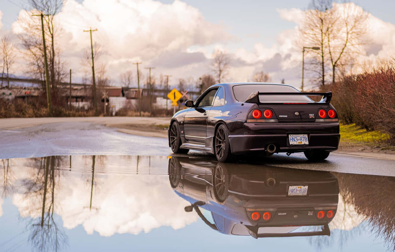 R33 GT-R 桌布 壁紙 — 一輛黑色跑車帶有紅色尾燈，停在一個水坑中，反射出黃昏時多雲的天空和光禿禿的樹木。