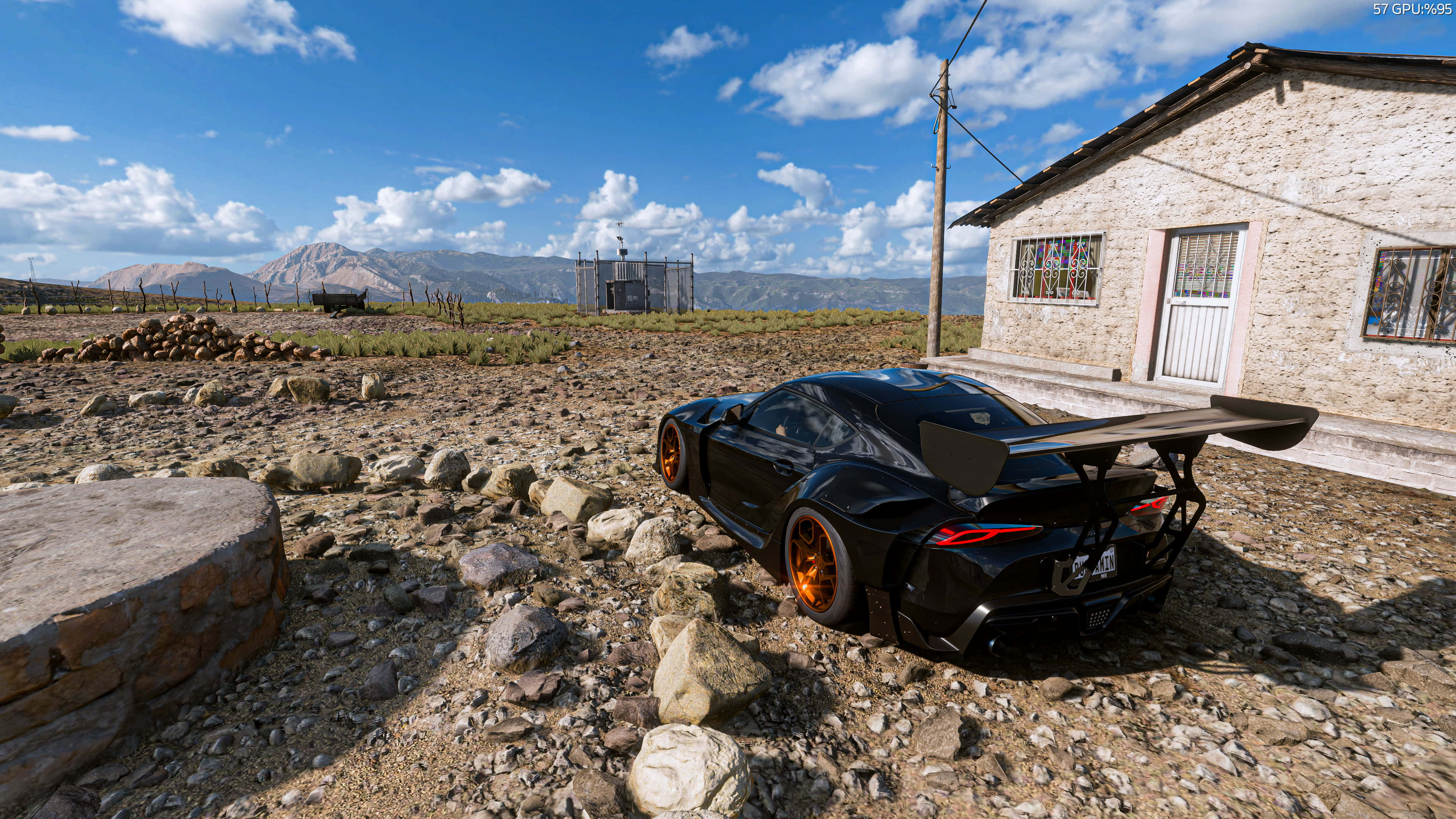 Forza Horizon 桌布 壁紙 — 一輛黑色跑車配橙色輪胎，停在石屋旁的岩石地形上，背景是明亮的藍天和遠山。