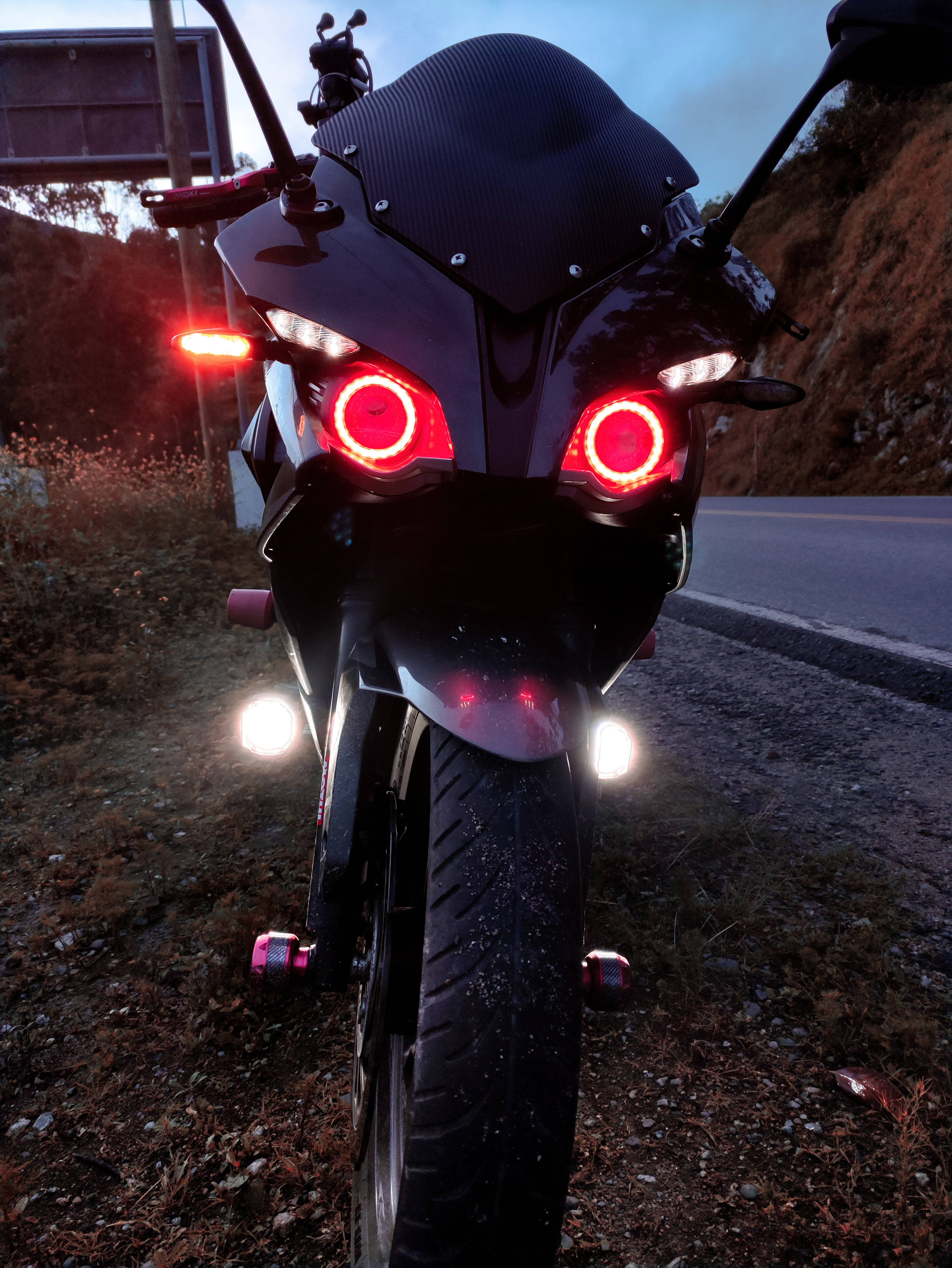 Pulsar 220 HD 桌布 壁紙 — 一輛黑色運動型摩托車，帶有發光的紅色圓形頭燈，在黃昏時停在路邊。