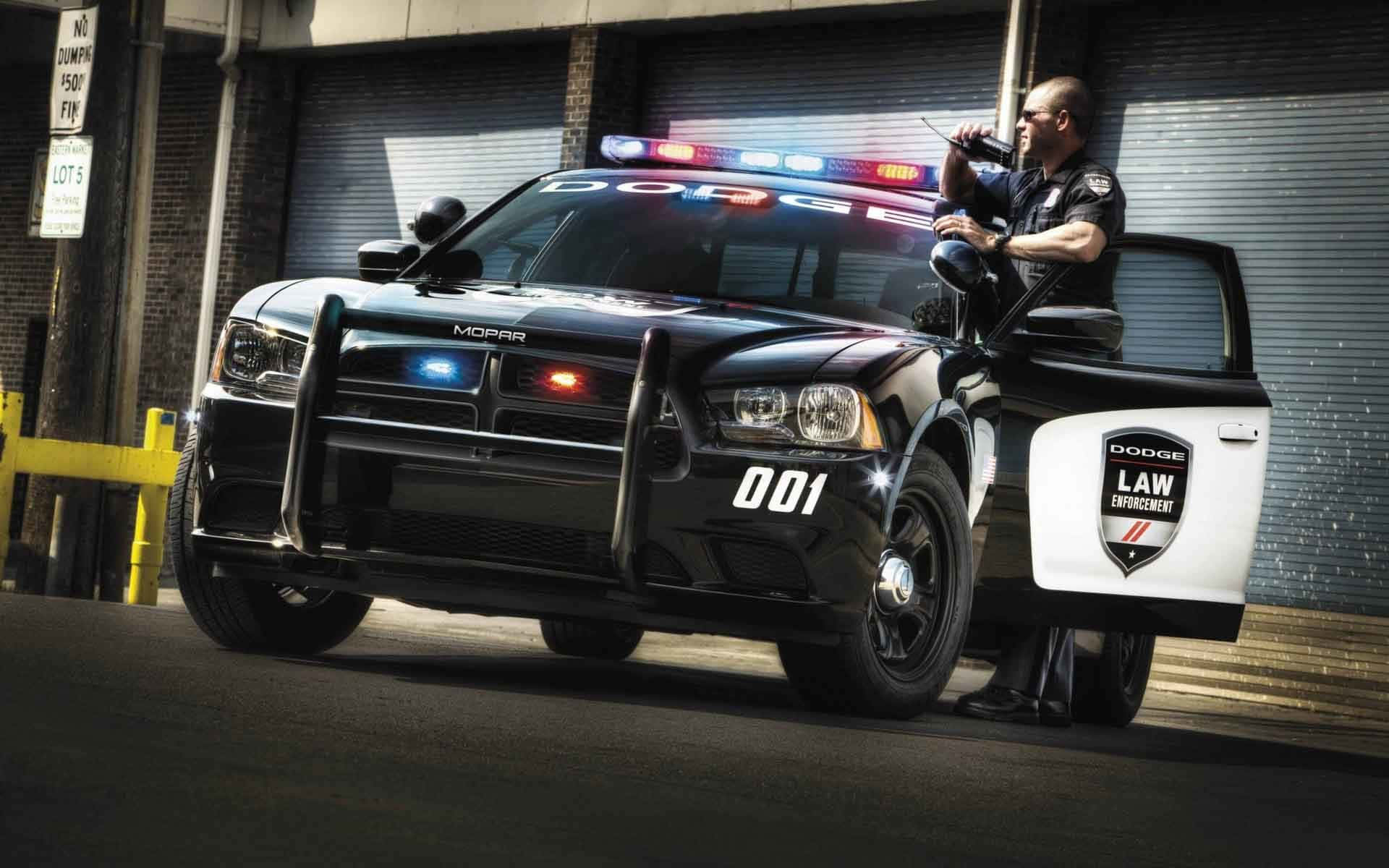Dodge 桌布 壁紙 — 一輛黑色道奇警車閃著燈停放在都市小巷中，一名警員探出車門，手持對講機。