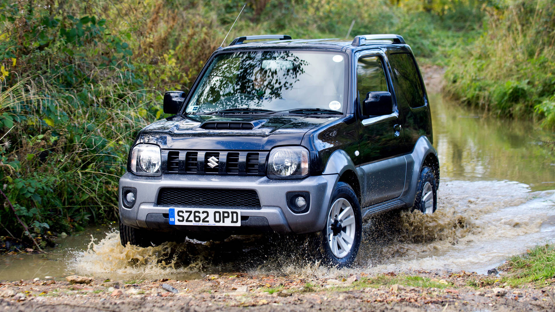 Suzuki 桌布 壁紙 — 一輛黑色鈴木 Jimny 在泥濘的森林溪流中濺起水花，秋葉在車輪周圍旋轉。