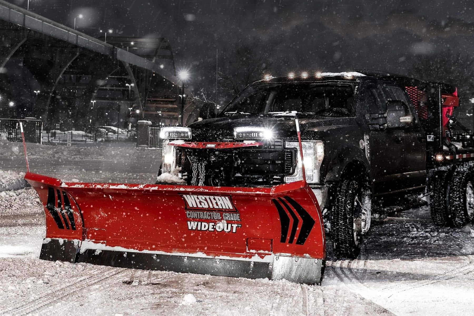 Snowplow 桌布 壁紙 — 一輛黑色除雪車帶有亮紅色鏟刀，在夜間街燈下的冬日城市環境中清除積雪。